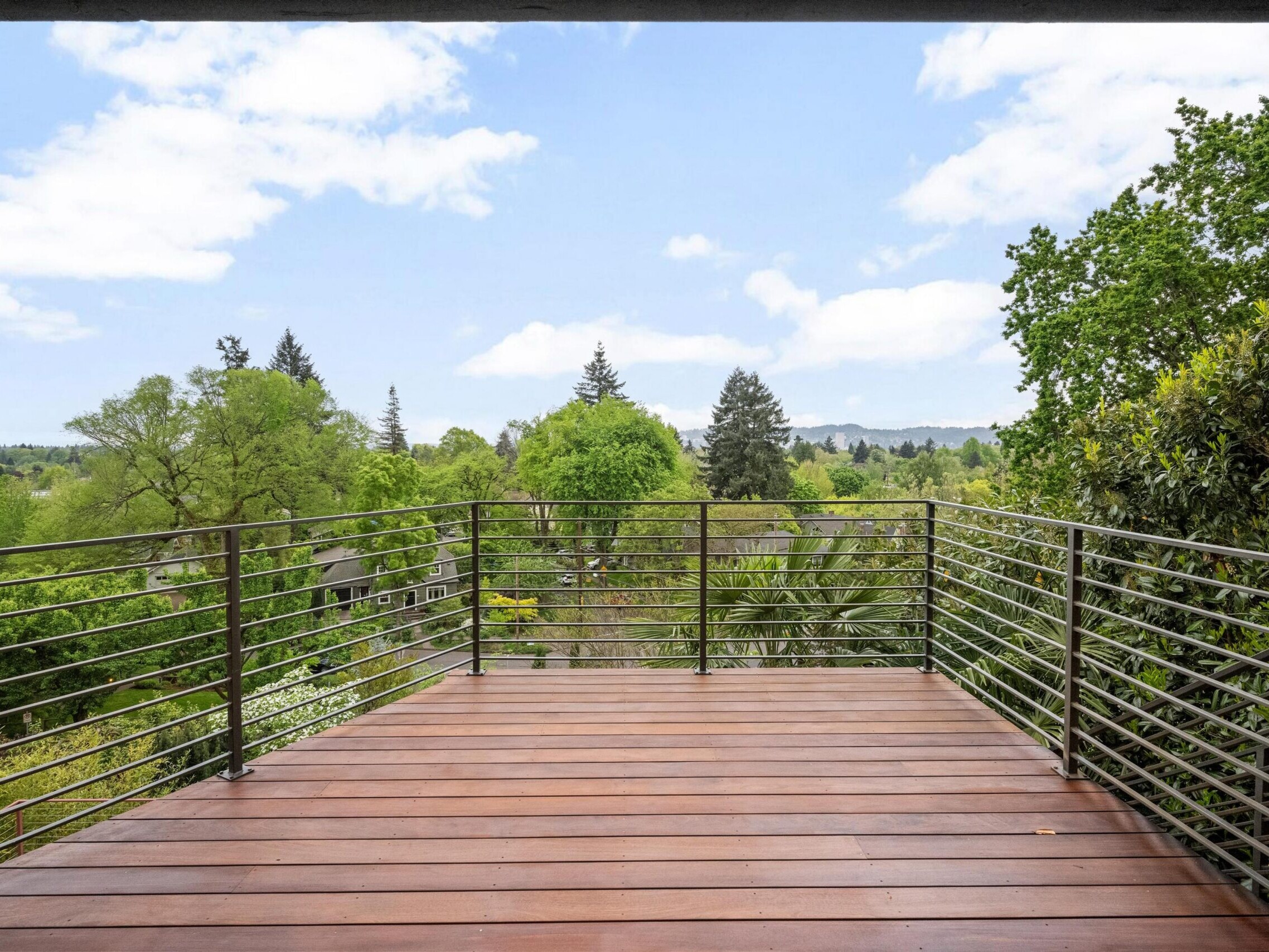A wooden deck with metal railings overlooks a lush, green landscape with trees and a partly cloudy sky in the background.