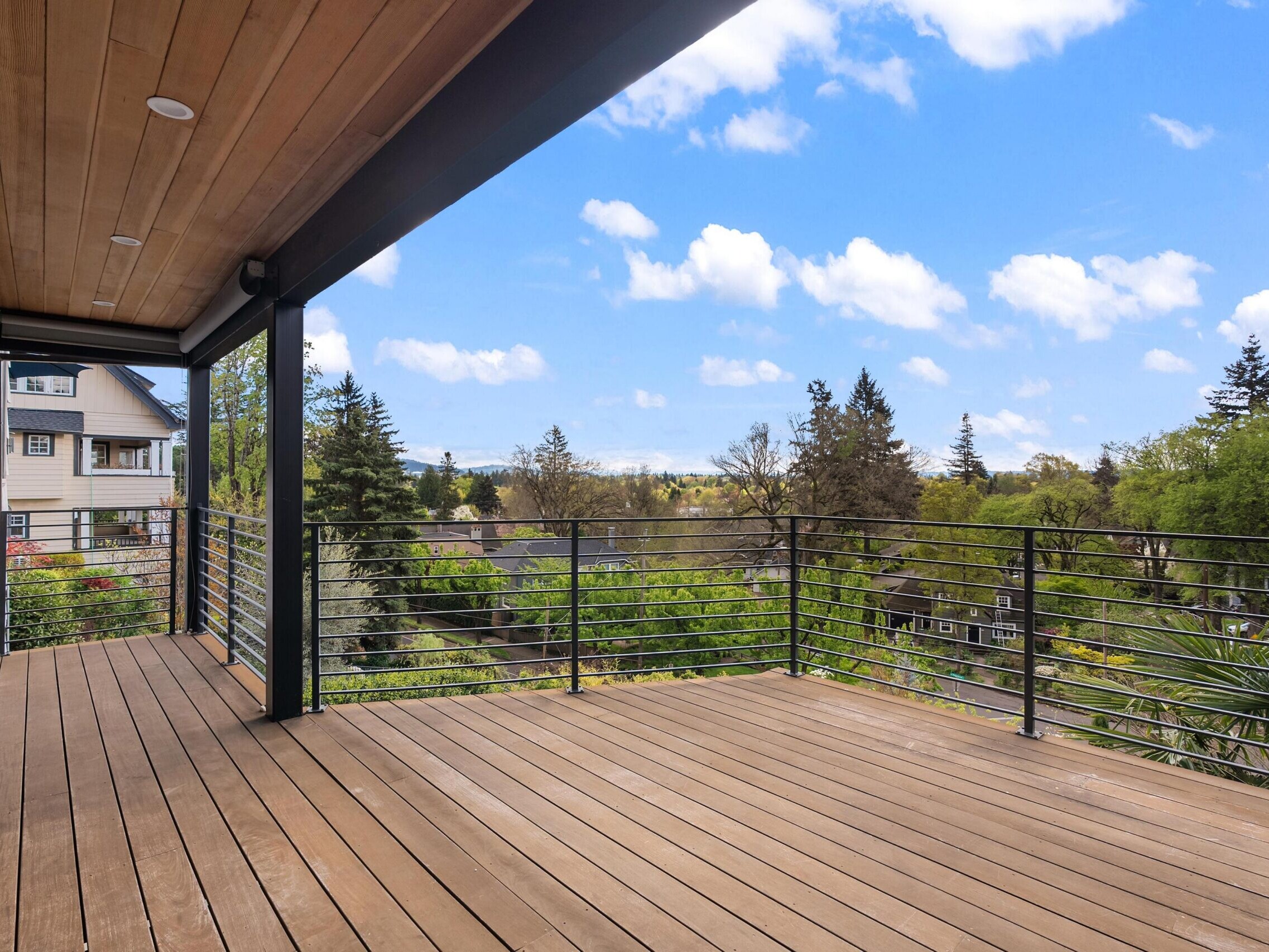 Spacious wooden deck with black metal railings, overlooking a scenic view of trees, houses, and a blue sky with scattered clouds.