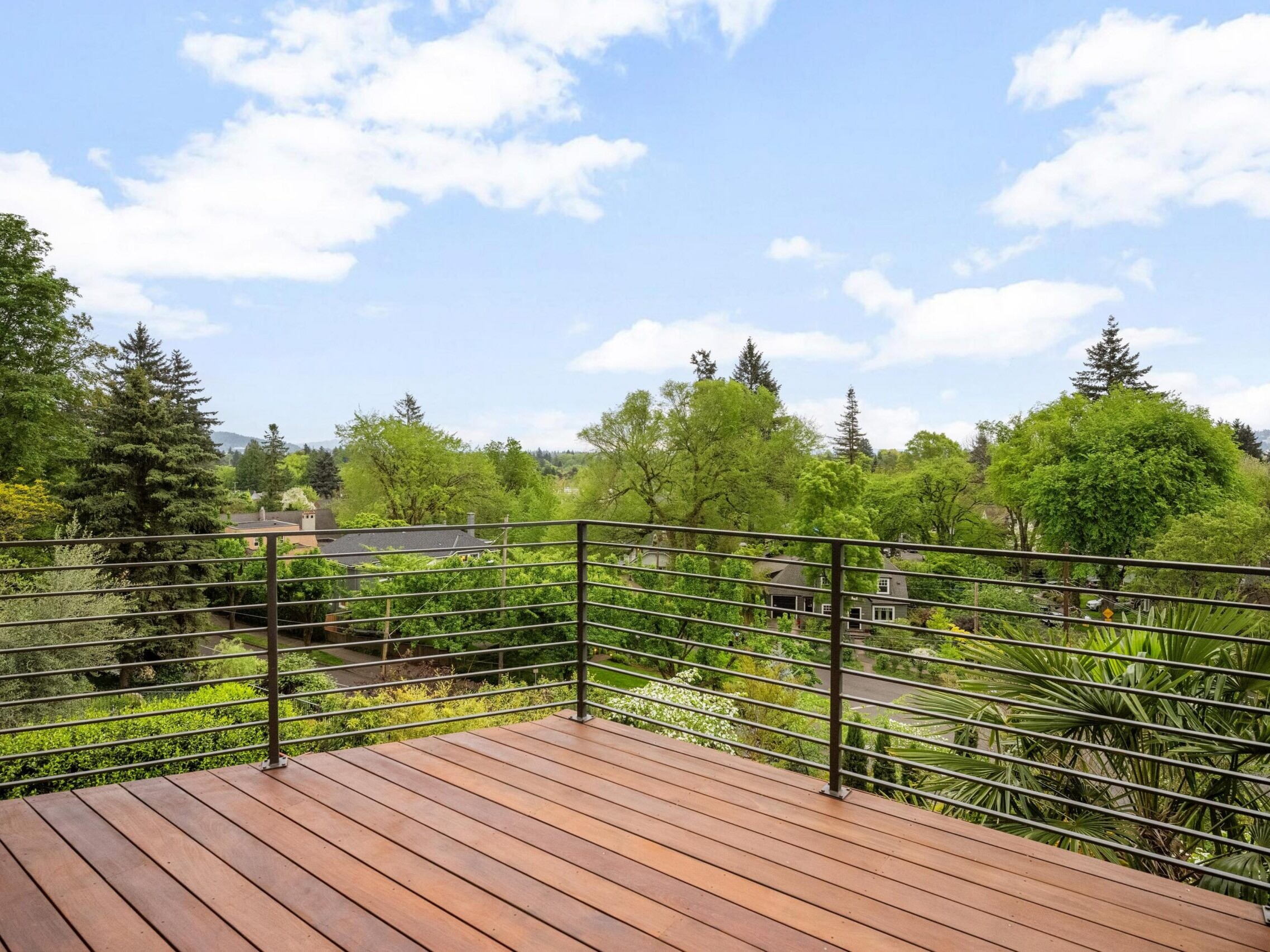 A wooden deck with a metal railing overlooks a lush, green neighborhood filled with trees under a partly cloudy sky.