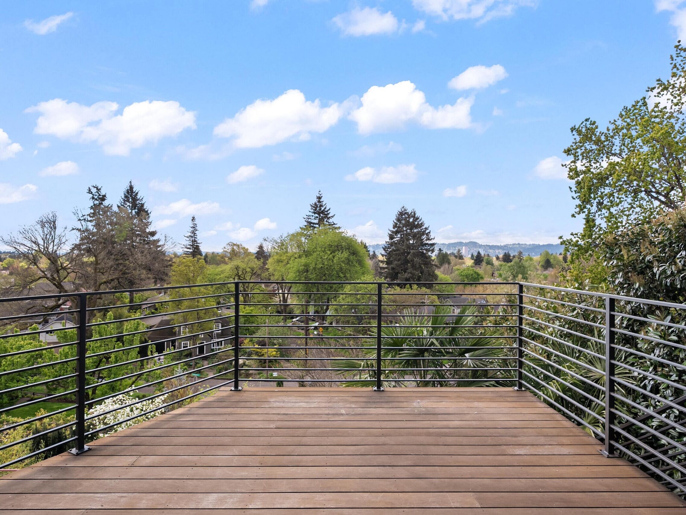 A wooden balcony with metal railings overlooks a lush, tree-filled neighborhood under a bright blue sky with scattered white clouds.