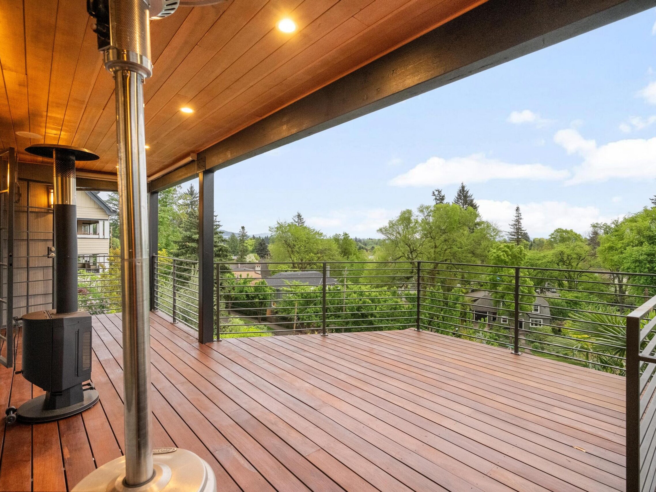 Covered wooden deck with metal railings, ceiling lights, and patio heaters, overlooking a lush, green suburban neighborhood and trees under a partly cloudy sky.