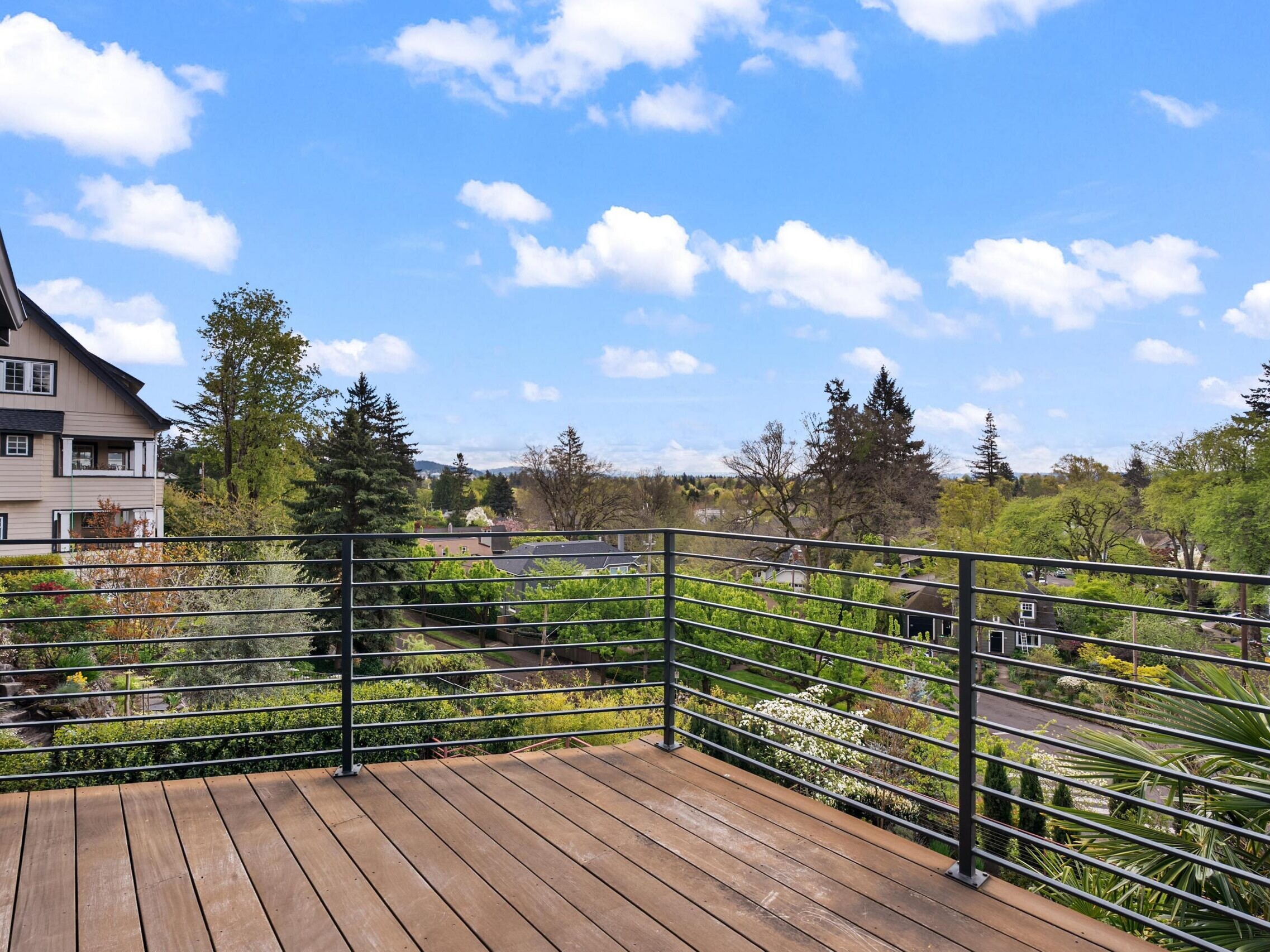 A wooden balcony with metal railings overlooks a neighborhood with trees, houses, and a clear blue sky with scattered clouds.