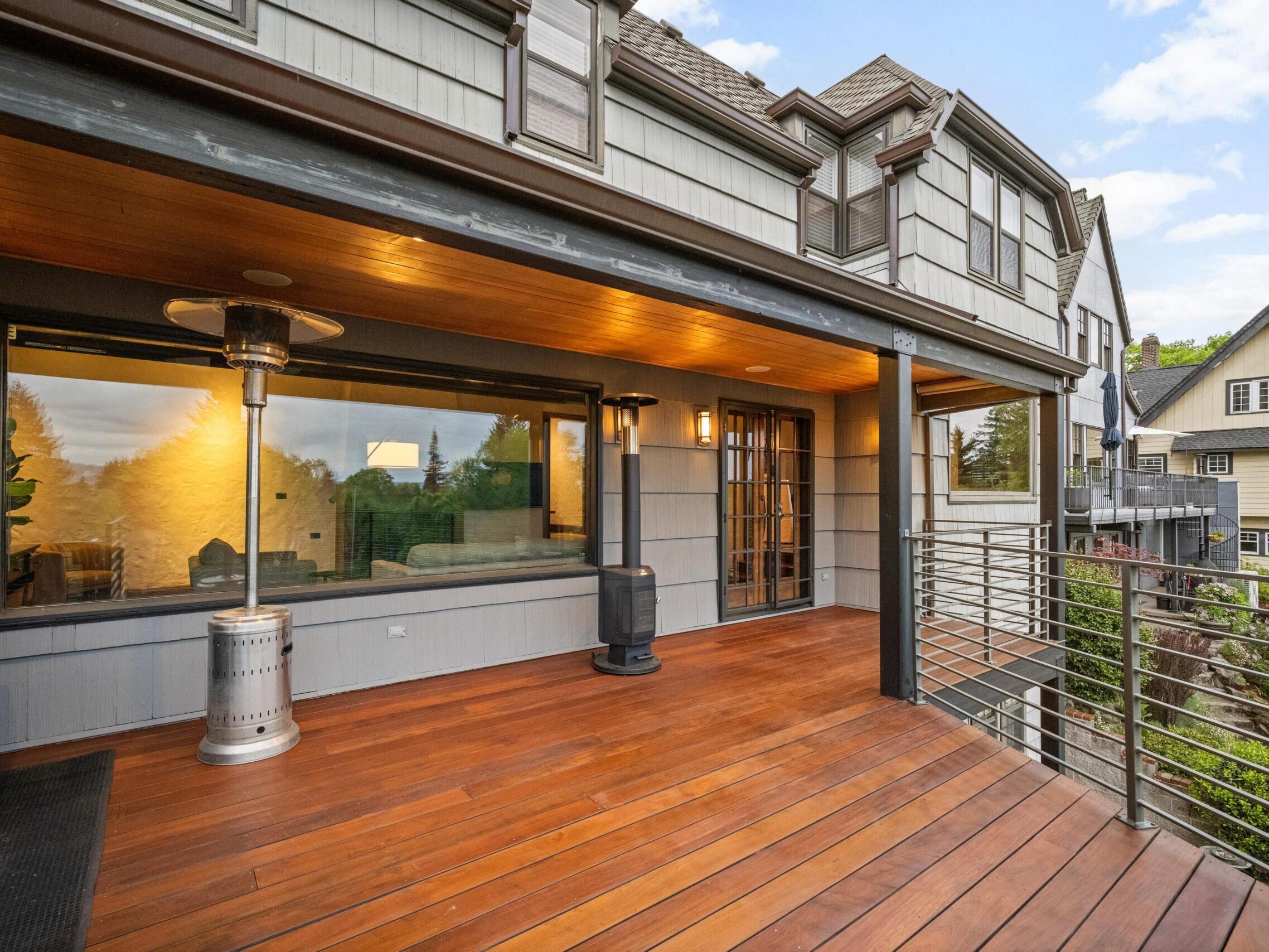 Spacious wooden balcony with metal railing, overhead lighting, and two outdoor heaters, attached to a modern house with large windows and surrounded by neighboring homes and greenery.