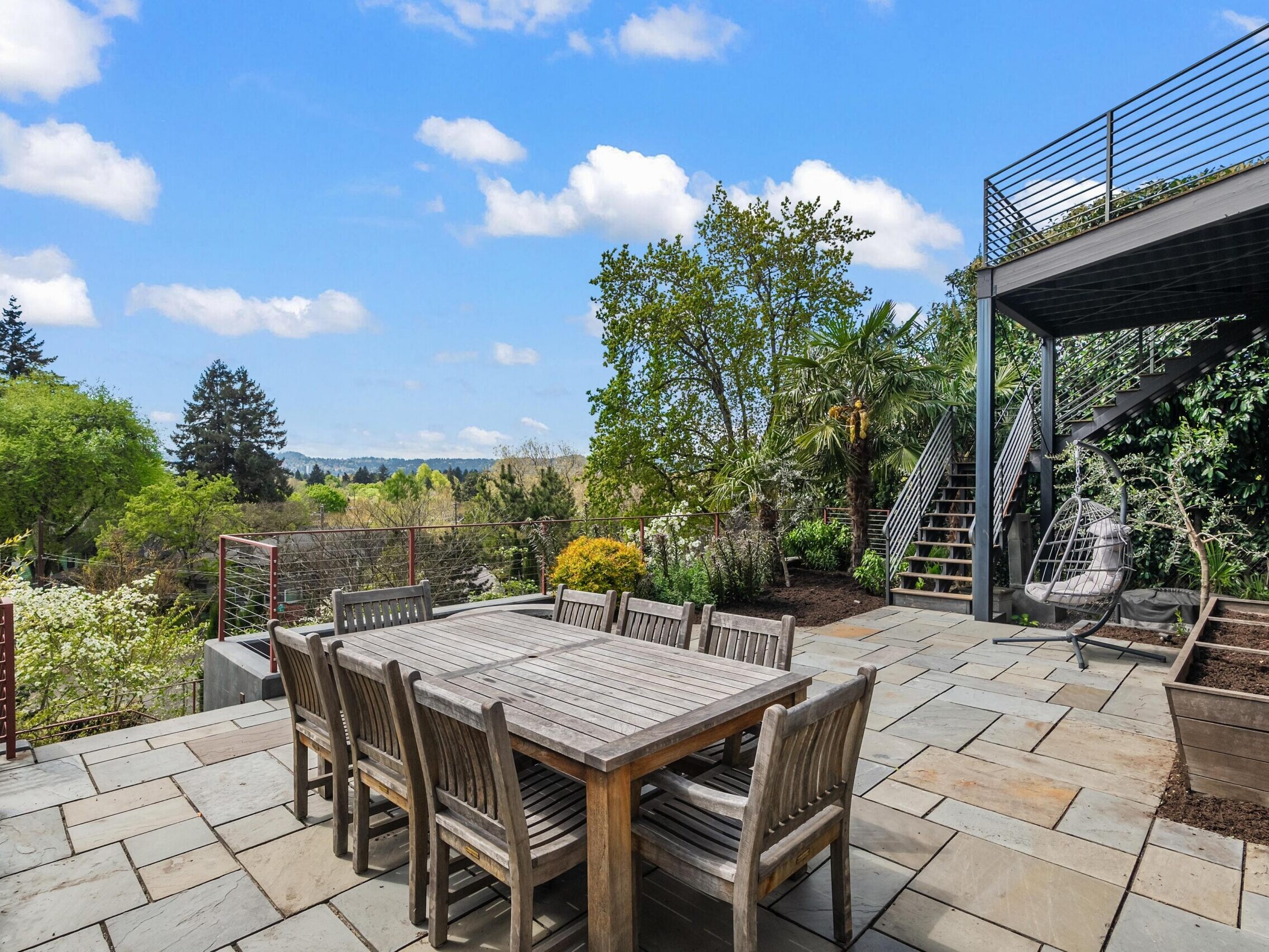 A spacious patio with a wooden dining table and chairs, surrounded by greenery. There is a metal staircase leading to an upper deck and a hanging chair in the corner. The sky is blue with scattered clouds.