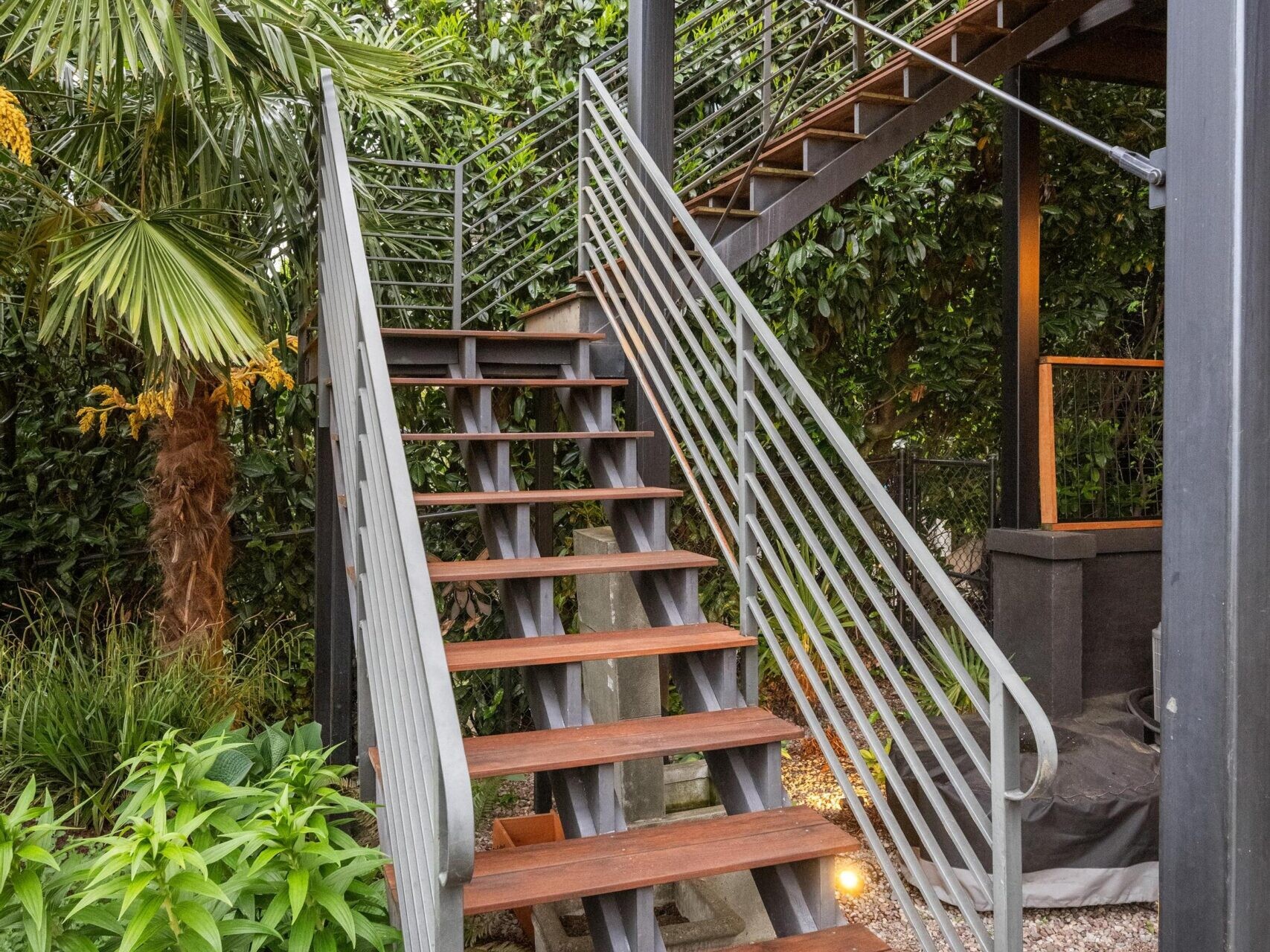 Outdoor staircase with wooden steps and metal railings leading up to a modern deck, surrounded by lush green plants and trees, with gravel and patio area below.