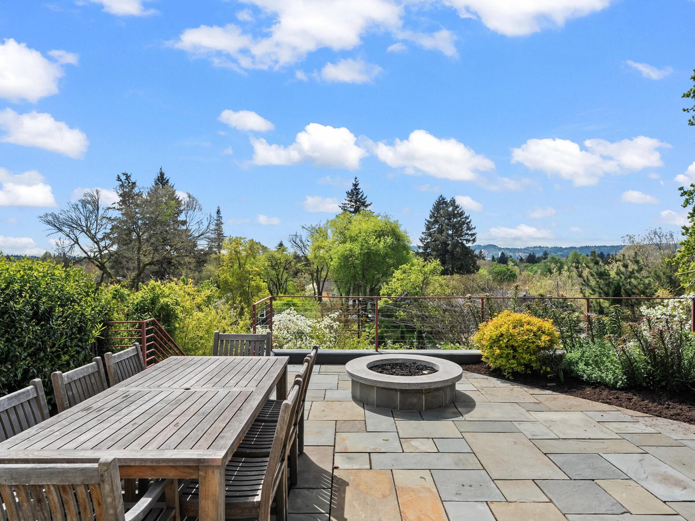 A spacious stone patio with a large wooden dining table and chairs, a circular fire pit, surrounded by lush greenery and trees, overlooking a scenic landscape under a blue sky with scattered clouds.