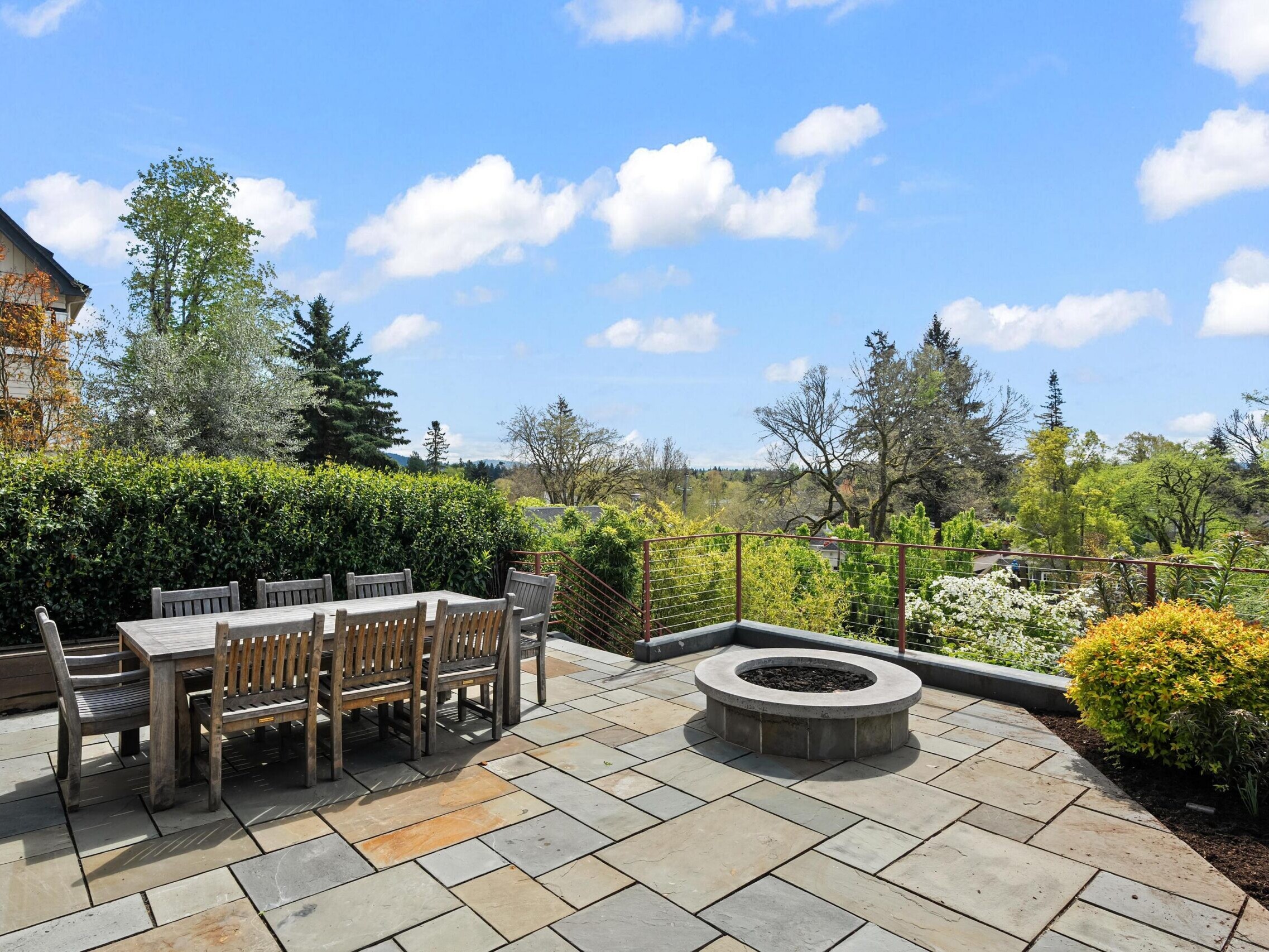 A spacious stone patio with a wooden outdoor dining table and chairs, a round fire pit, and lush greenery, overlooking a scenic view of trees and a blue sky with clouds.