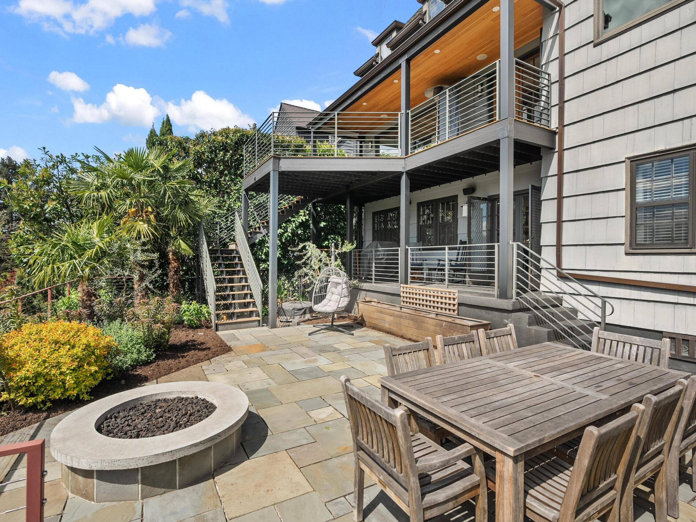 Spacious backyard patio with a wooden dining table, fire pit, garden area, and two-story deck with stairs, under a partly cloudy sky beside a modern house.