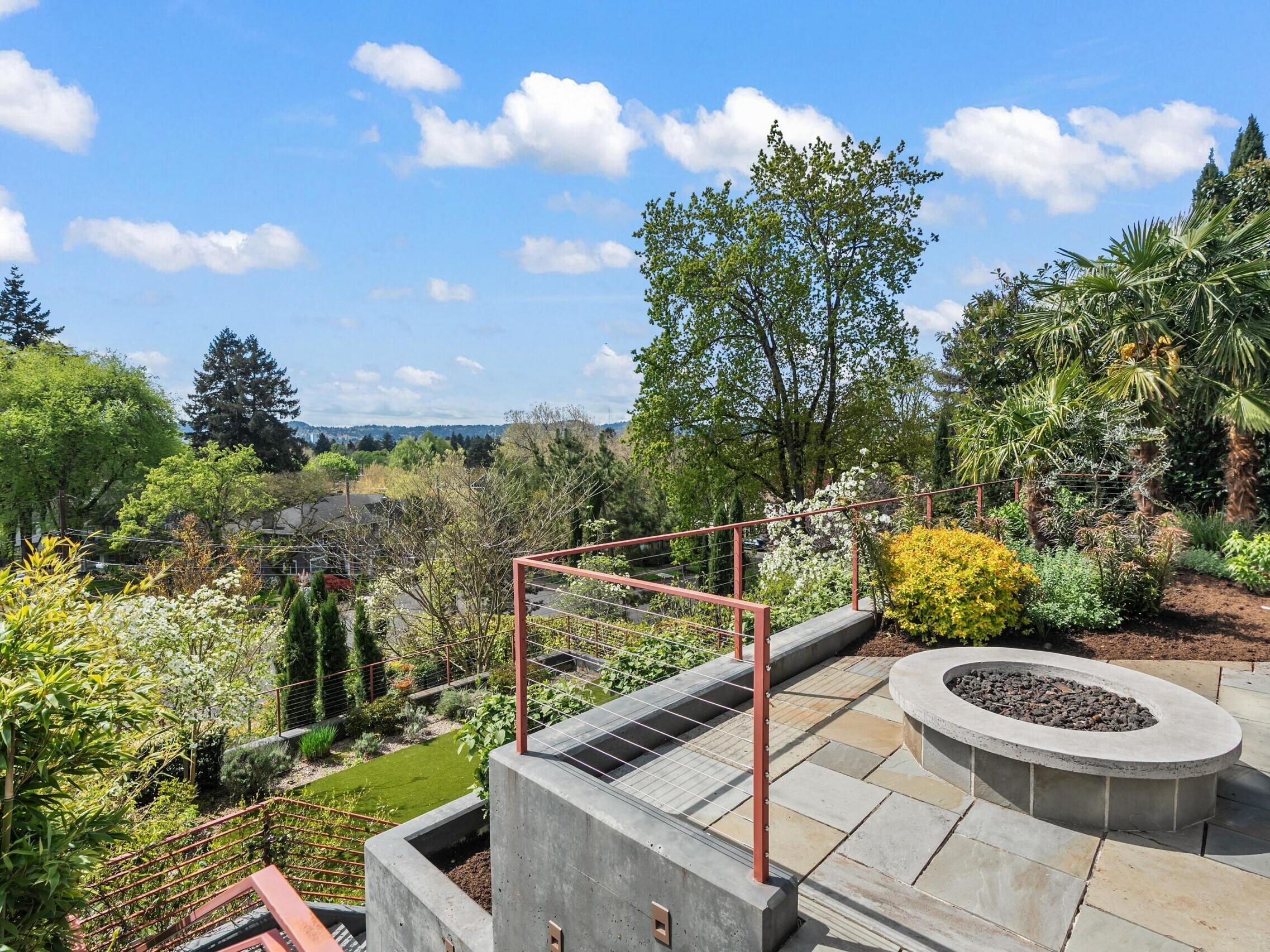Stone patio with circular fire pit, metal railing, and steps overlooking a lush, landscaped garden and distant trees under a blue sky with scattered clouds.