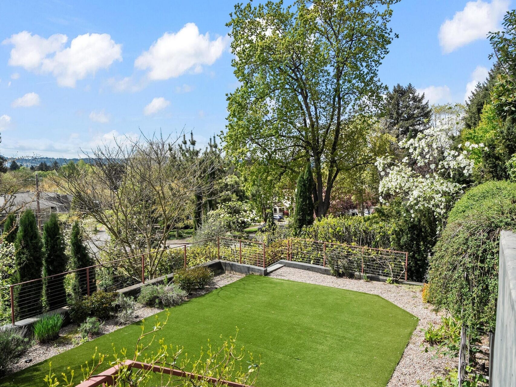 View from the top of outdoor steps overlooking a green lawn, gravel pathways, lush trees, and landscaped gardens under a bright blue sky with scattered clouds.