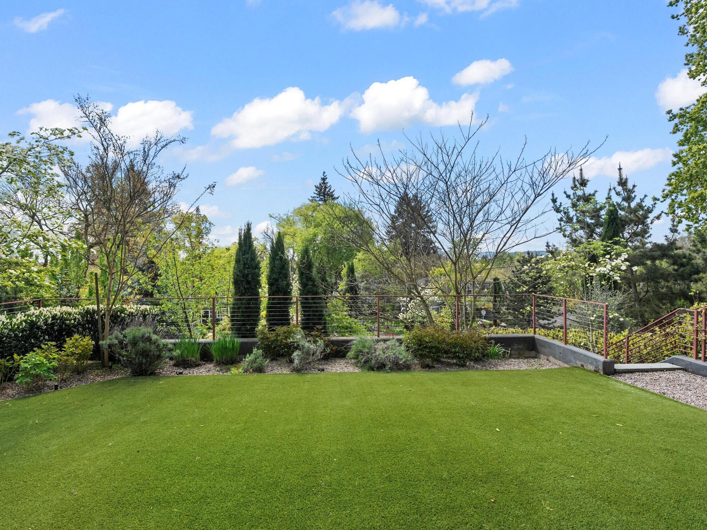 A well-maintained backyard with a lush green lawn, various shrubs and small trees along the edges, a gravel path, and a metal railing, all under a bright blue sky with scattered clouds.