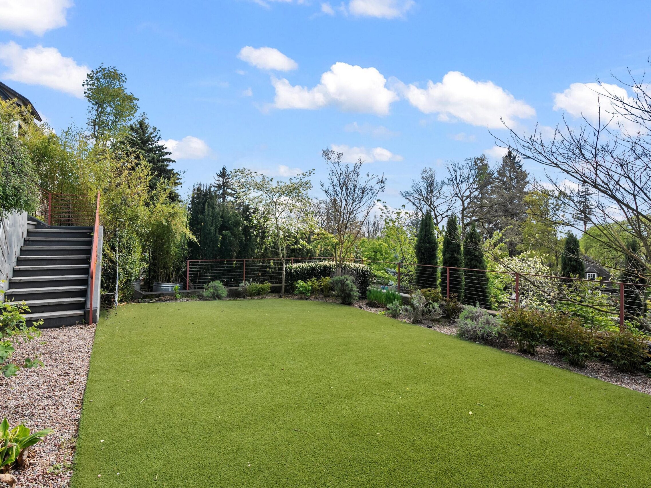 A landscaped backyard with artificial grass, gravel borders, stairs on the left, and various trees and shrubs under a blue sky with scattered clouds.