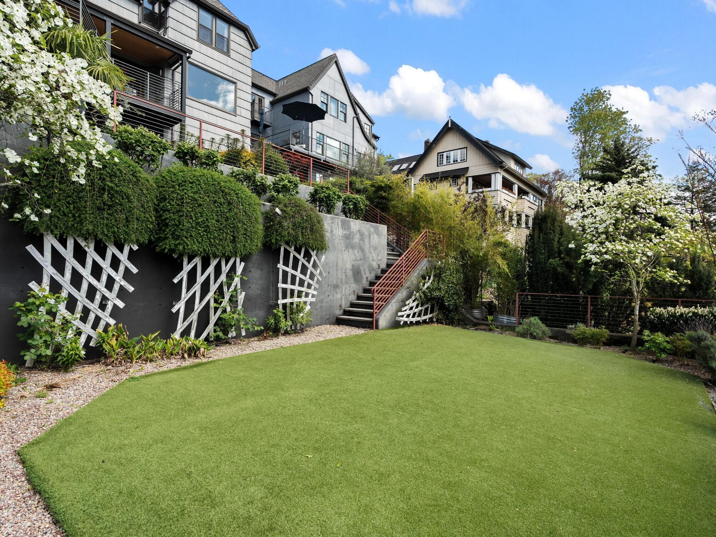 A modern backyard with a manicured green lawn, gravel paths, landscaped plants, trellises on a retaining wall, a stairway, and contemporary houses in the background under a blue sky with clouds.