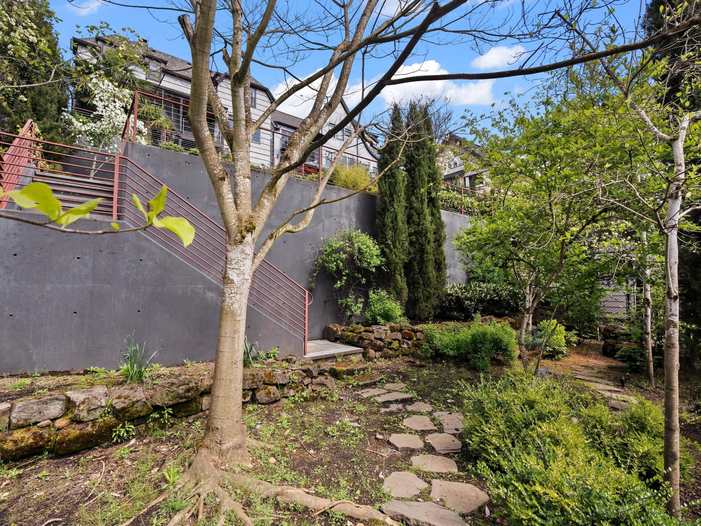 A landscaped backyard with a stone pathway, green trees, shrubs, and a gray retaining wall with a red metal railing. Steps lead up to a patio area, and houses are visible in the background under a blue sky.