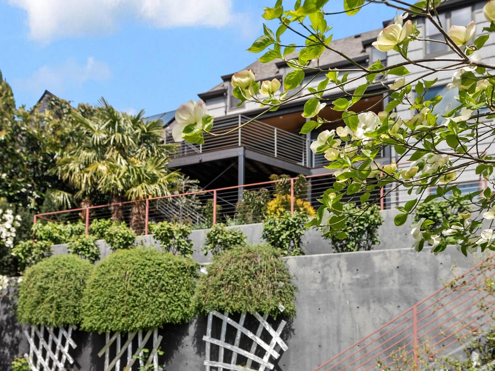 A modern building with a terrace overlooks a landscaped garden with green grass, climbing plants on trellises, and lush trees under a blue sky with white clouds.