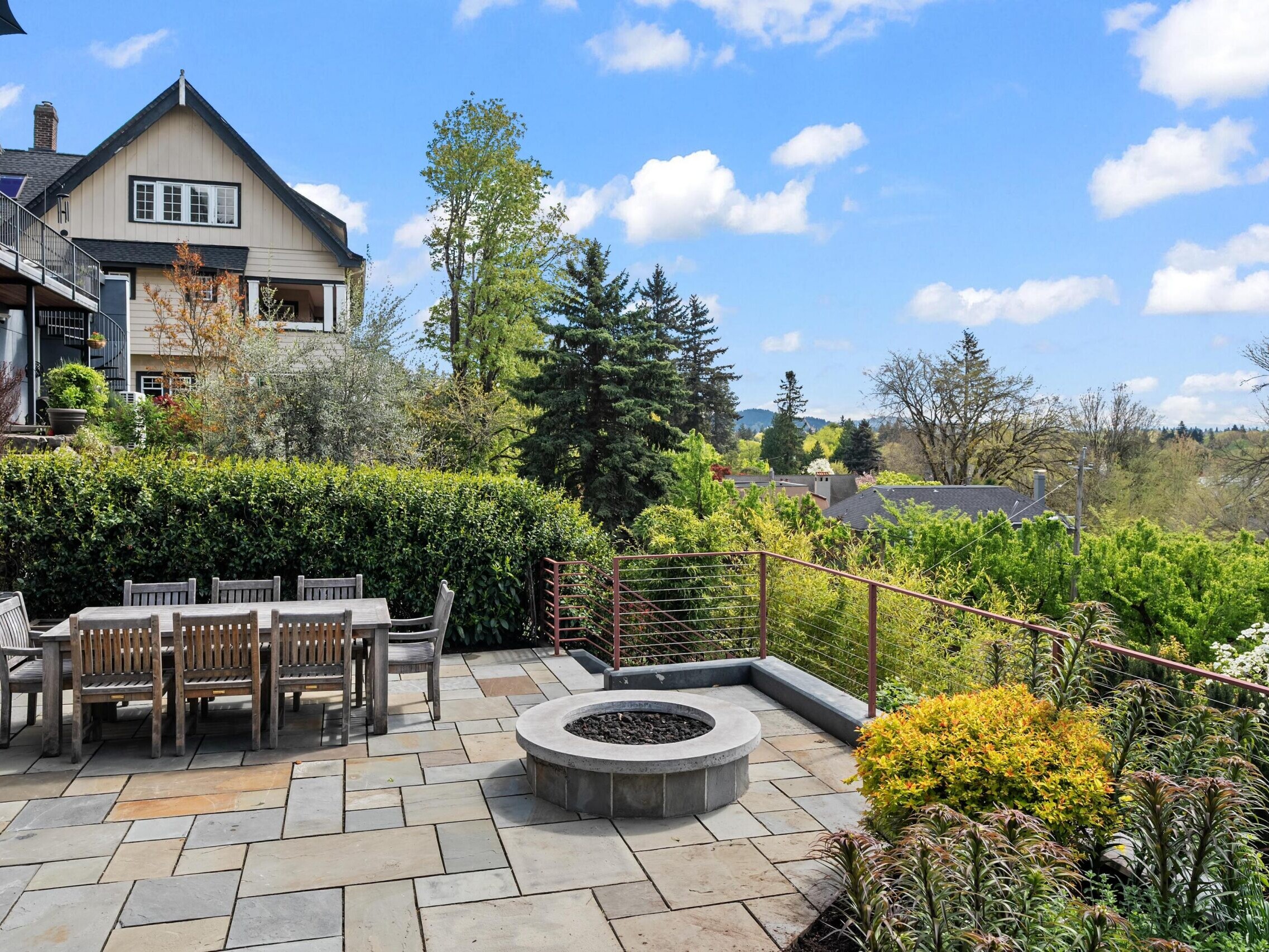 Spacious stone patio with a wooden dining table, chairs, and a circular fire pit, surrounded by lush greenery and garden beds, overlooking a scenic neighborhood with trees and houses under a blue sky.