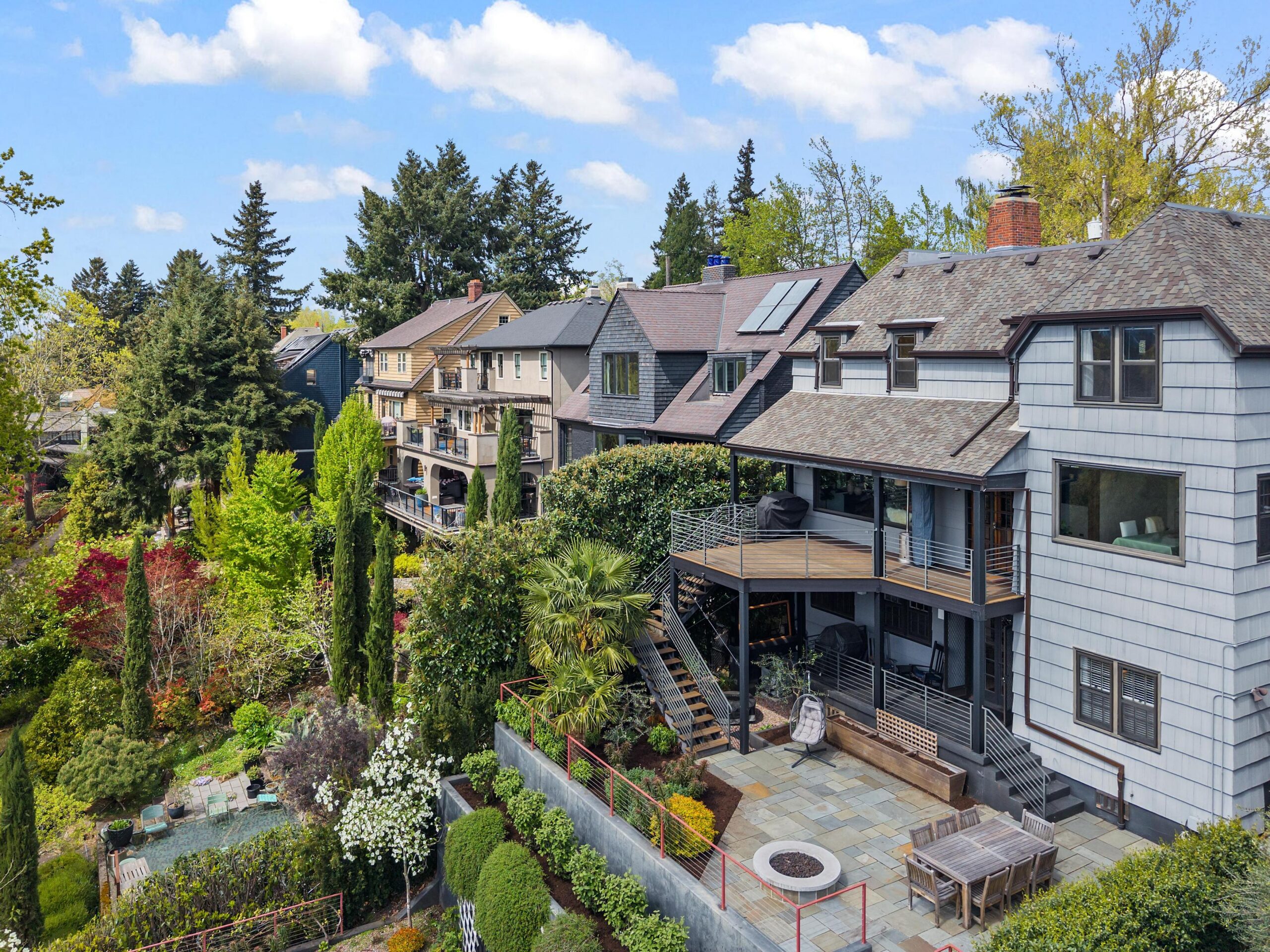 A row of multi-story homes with decks and landscaped yards on a hillside, featuring lush greenery, trees, a patio with a fire pit, and a partly cloudy sky above.