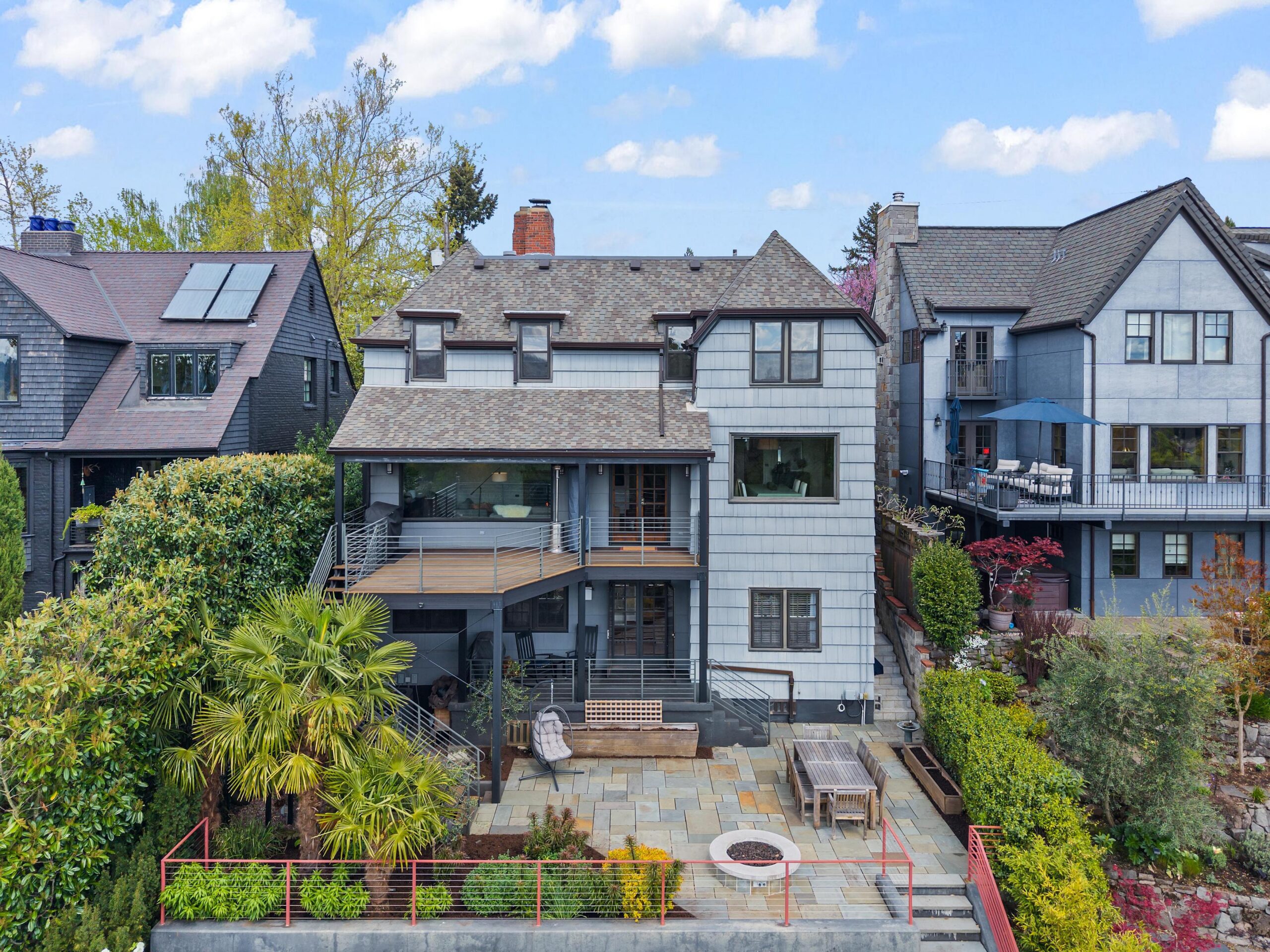 A large, multi-story gray house with balconies, patio seating, a fire pit, and landscaped greenery, situated between two other similar style homes on a sunny day.