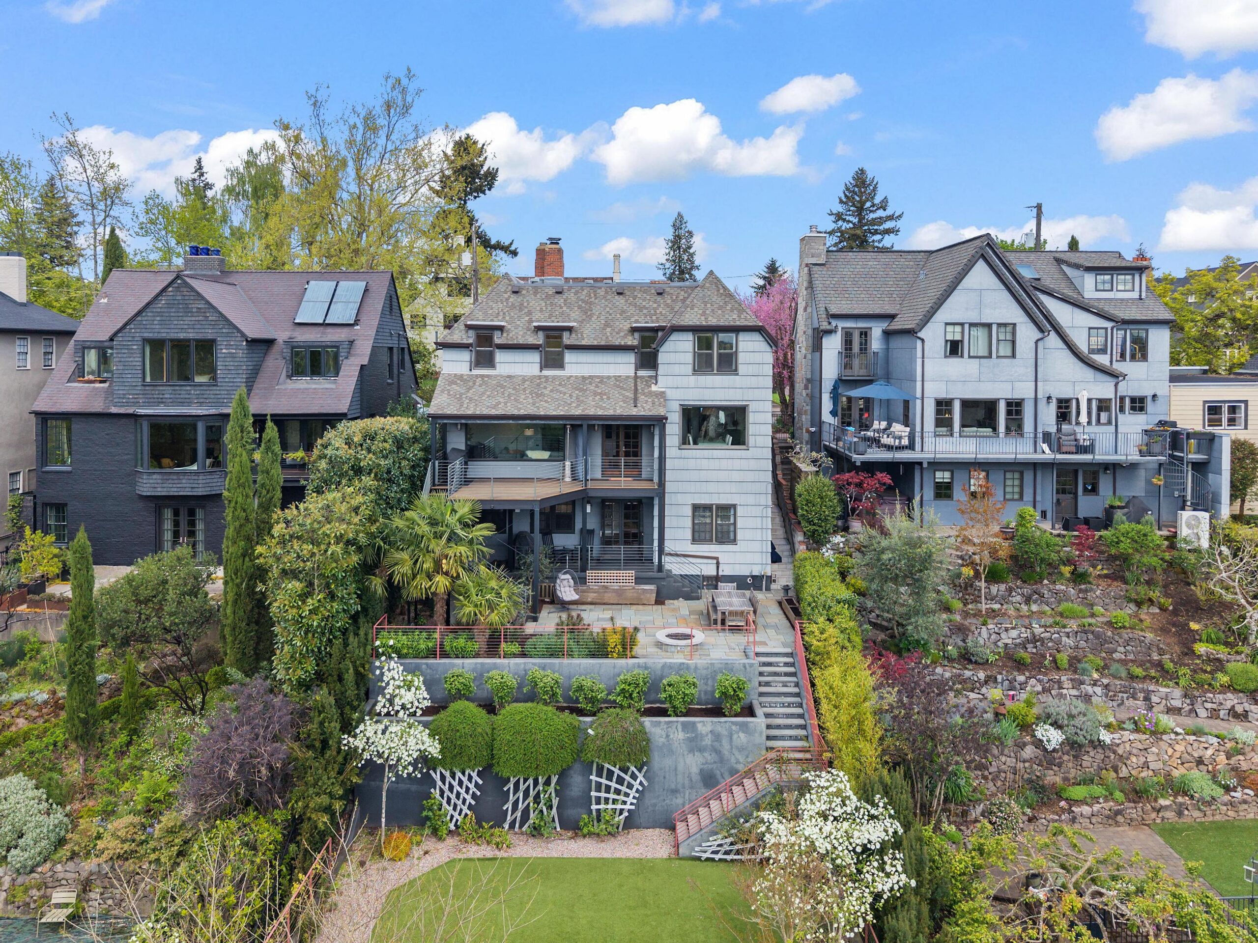 Three multi-story houses with gabled roofs overlook a landscaped garden with stone walls, shrubs, and a patio. Trees and flowering plants surround the homes under a bright blue sky with scattered clouds.