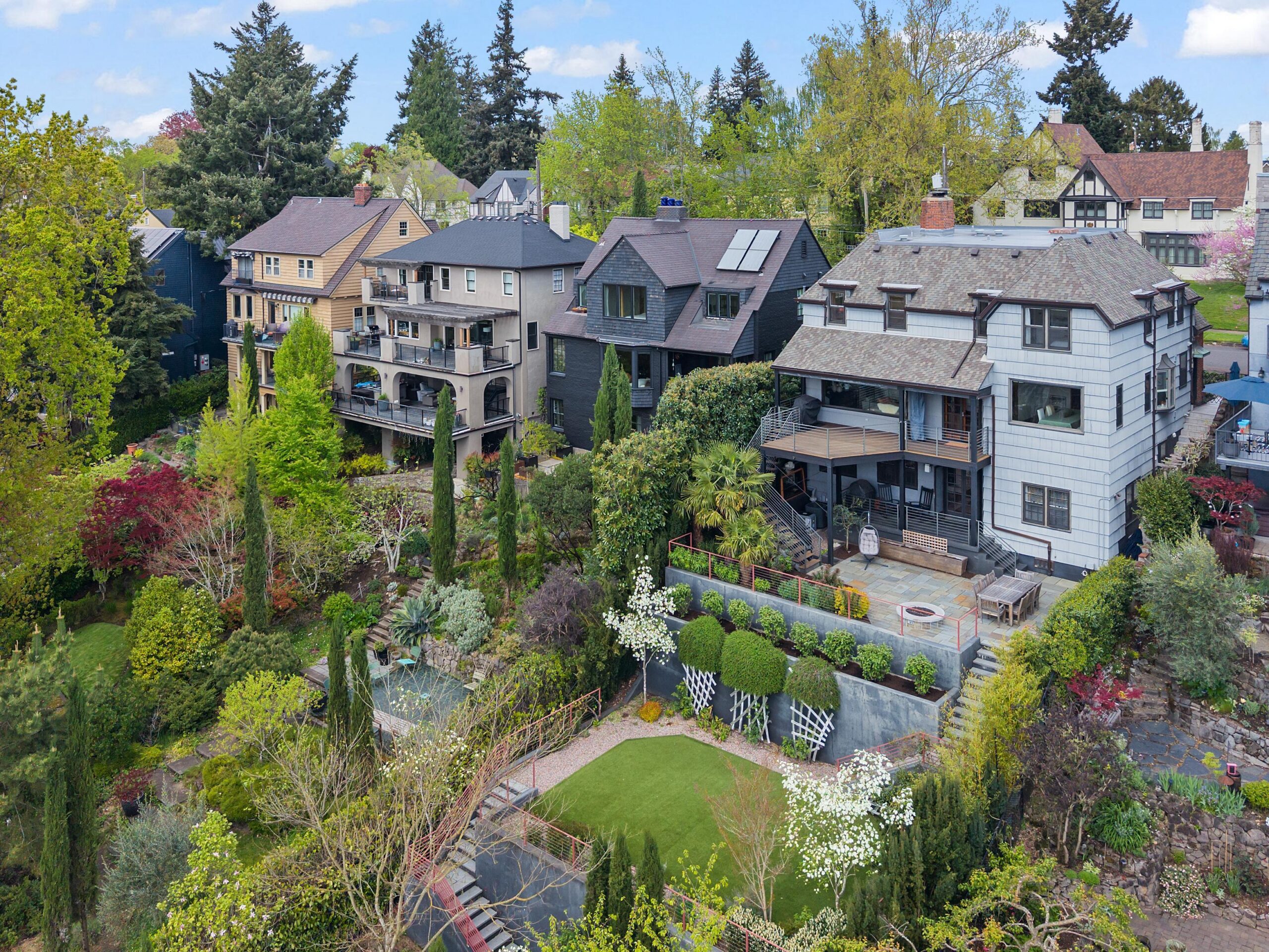 Aerial view of several large, multi-story homes with balconies, surrounded by lush gardens, manicured lawns, and tall trees in a residential neighborhood.