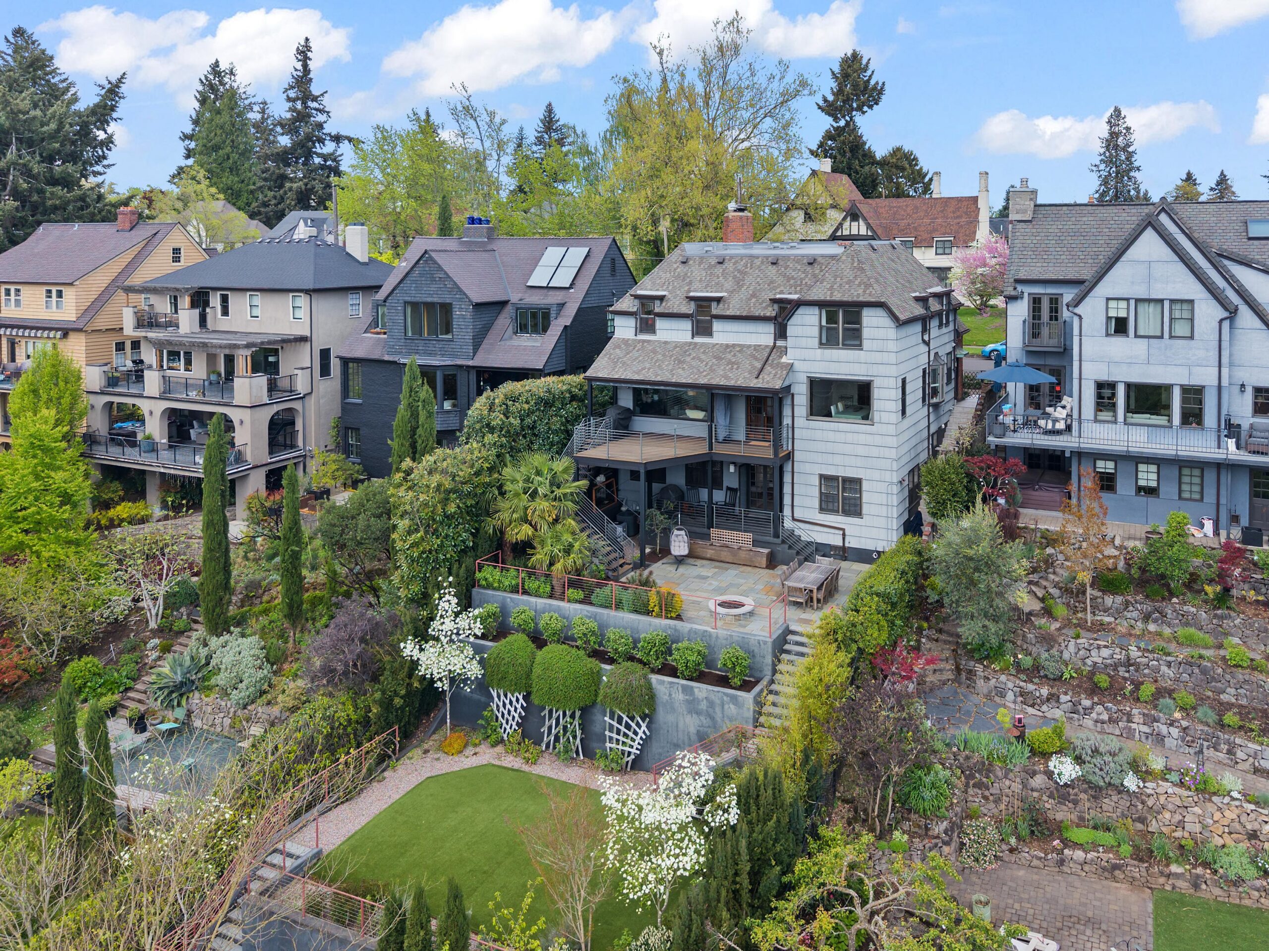 A row of large, multi-story houses with balconies and gardens sits on a hill, surrounded by lush greenery, trees, and landscaped yards under a blue sky with scattered clouds.