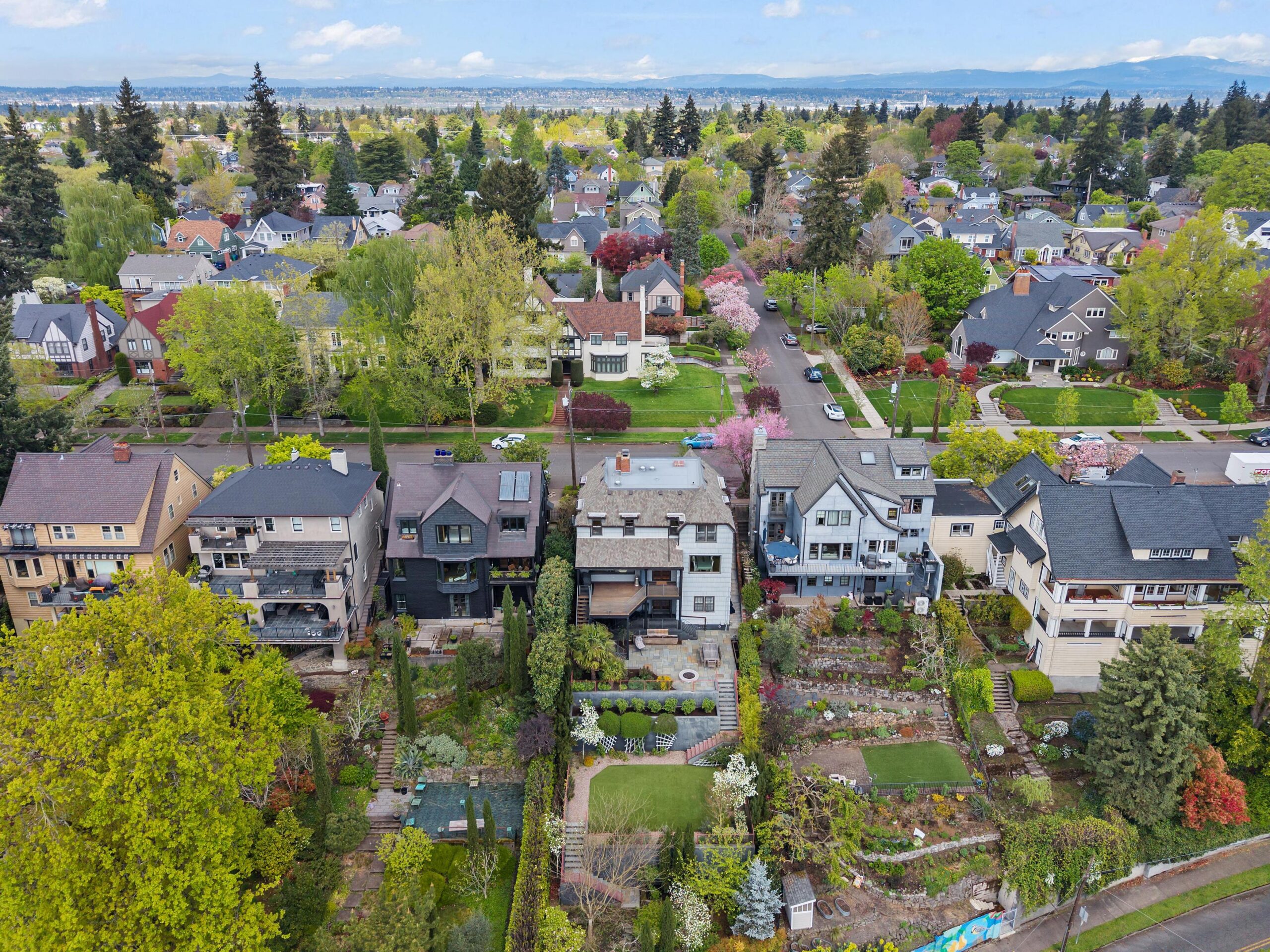 Aerial view of a residential neighborhood with rows of houses, lush green trees, and landscaped gardens. Streets run between the houses, and the background shows more houses and distant hills under a partly cloudy sky.
