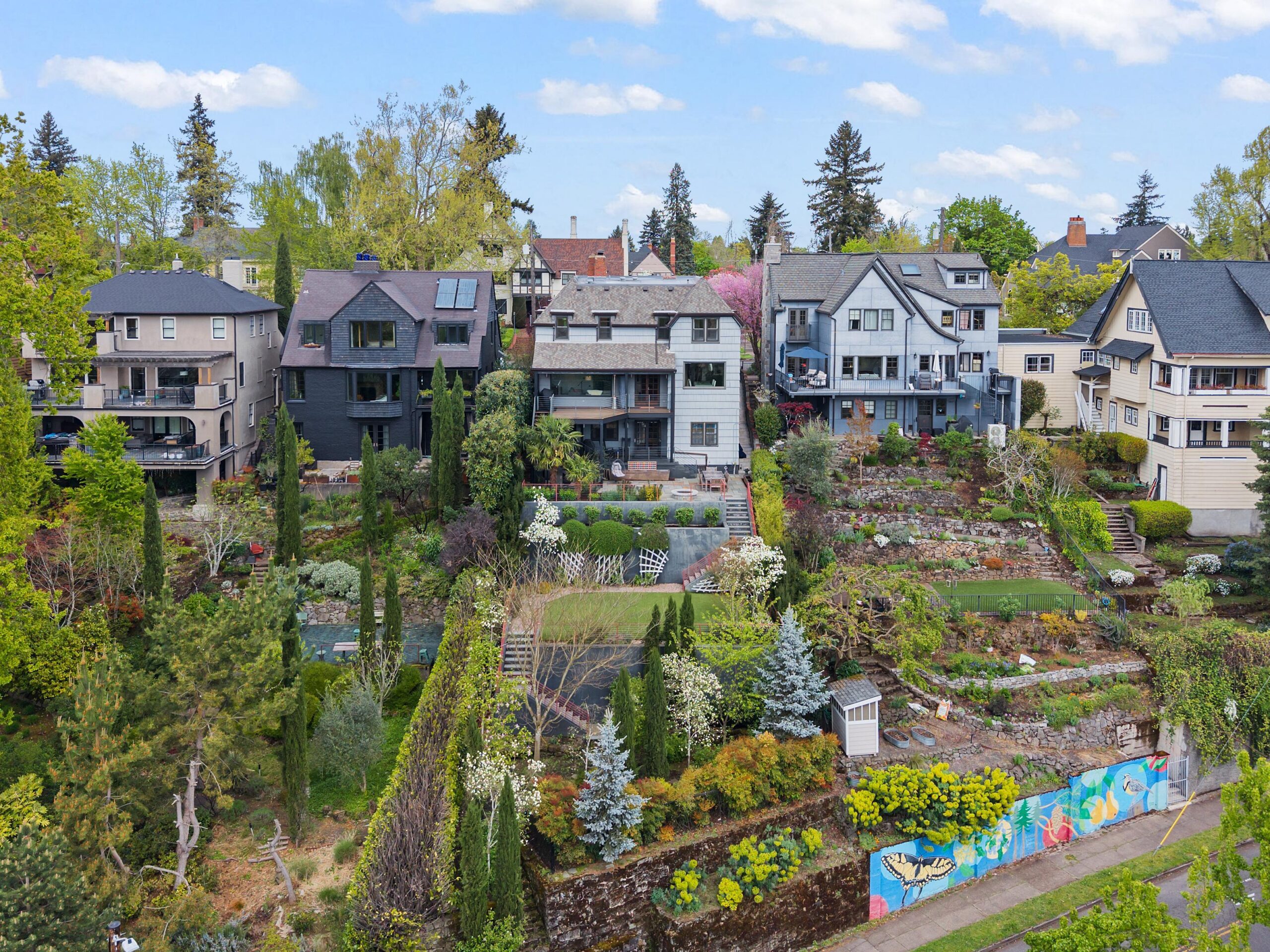 A row of large, multi-story houses with colorful gardens and terraced landscaping on a hillside. A mural decorates a retaining wall at the bottom, and tall trees frame the area under a blue sky with scattered clouds.