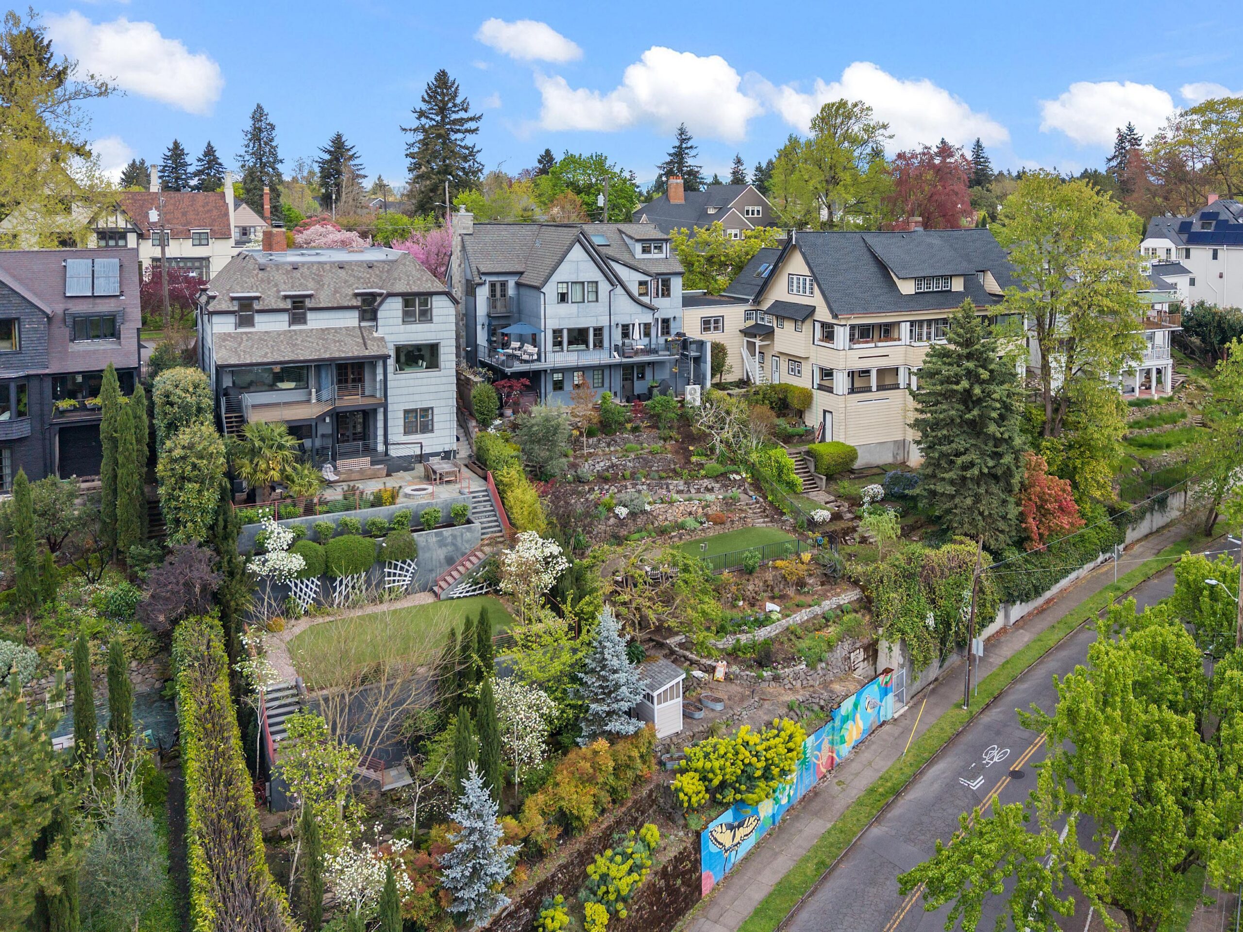 A row of large, multi-story houses with landscaped gardens on a hillside, overlooking a street with a colorful mural along the retaining wall. Trees and greenery surround the homes under a blue sky with clouds.