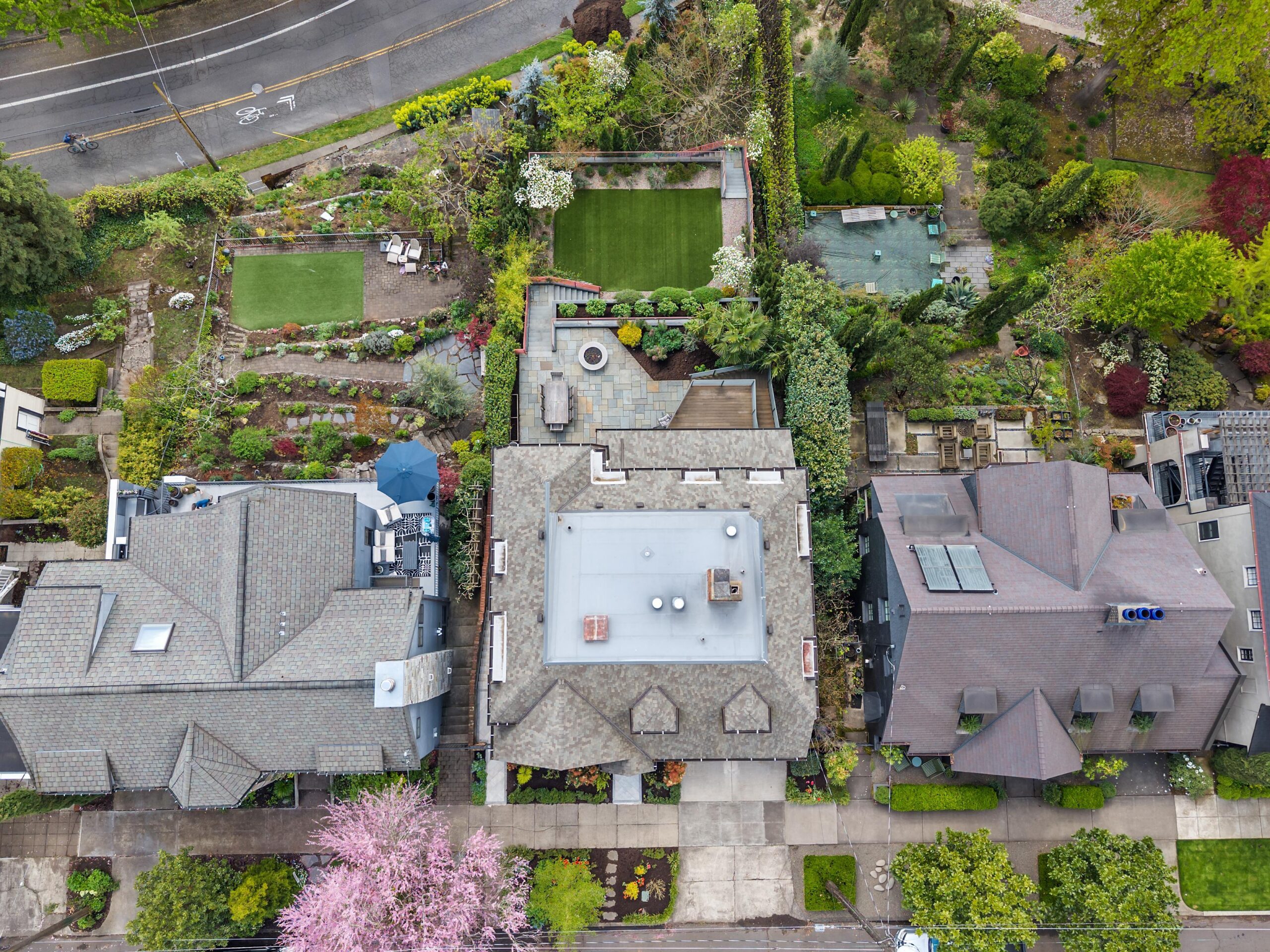 Aerial view of three houses lined up side by side with gardens, lawns, trees, and a street in the background. The roofs and yards show landscaping details and outdoor seating areas.