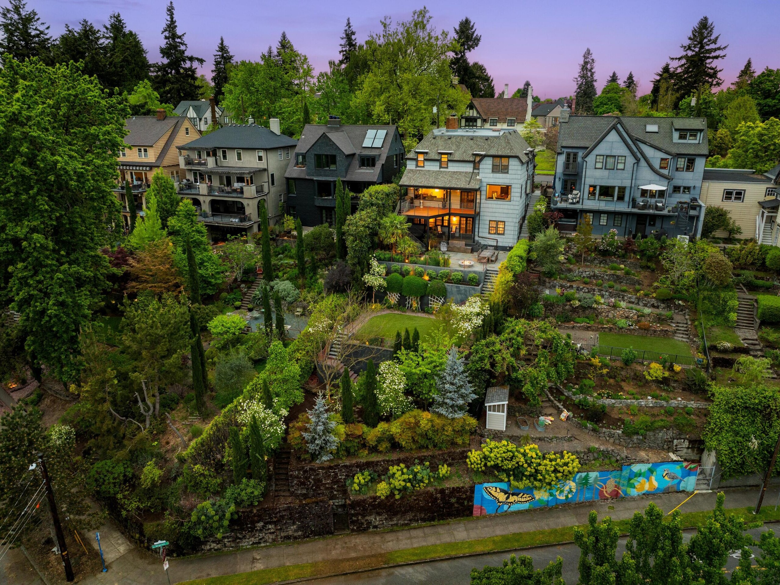 Aerial view of a neighborhood at dusk with large homes surrounded by lush gardens and trees. One house is brightly lit, and a colorful mural decorates a retaining wall along the street.