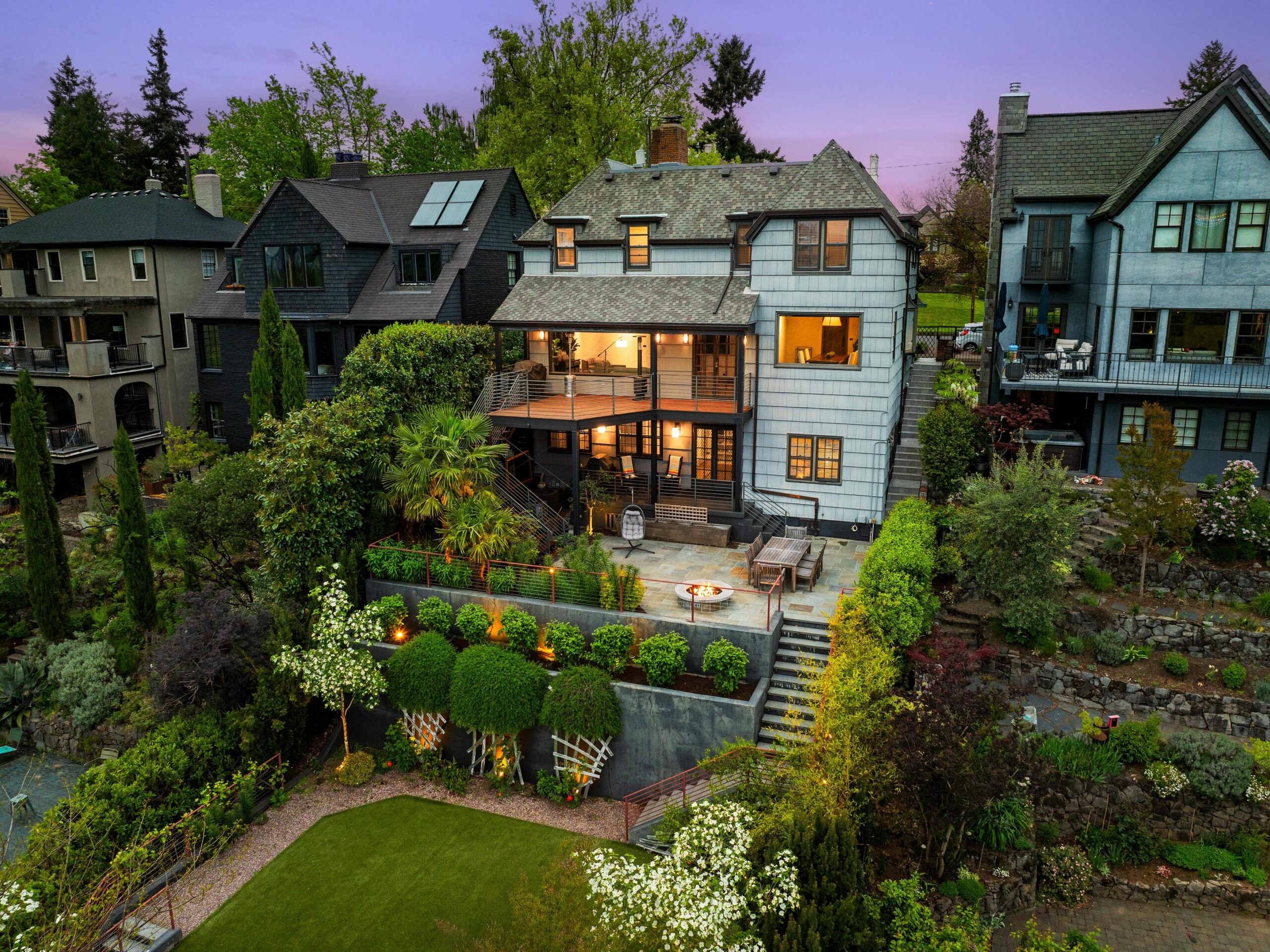 A large, elegant house with multiple stories and balconies is surrounded by lush gardens, trees, and a manicured lawn, with neighboring homes close by and a purple evening sky in the background.