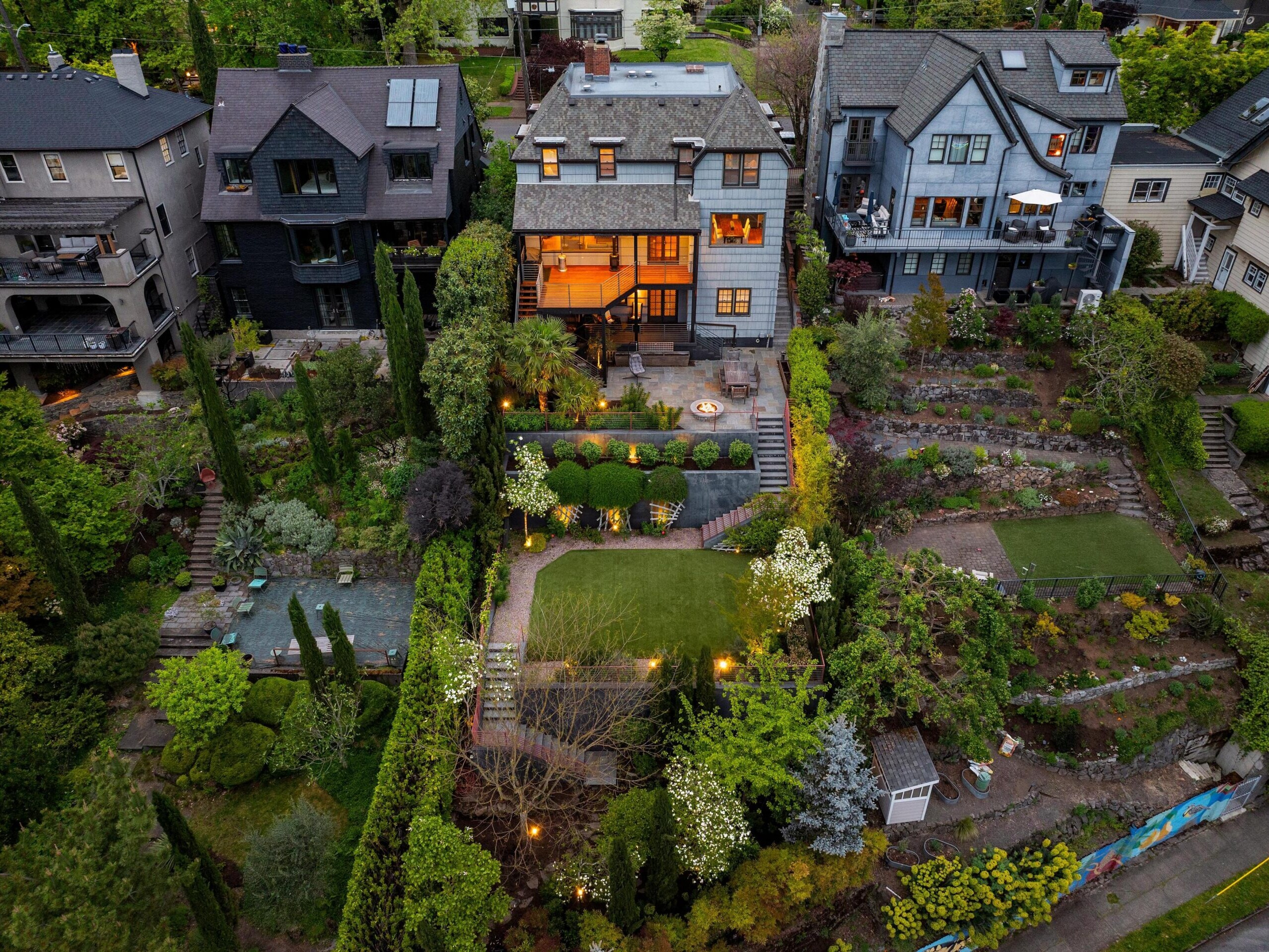 Aerial view of a large, modern house with a lit-up patio, surrounded by lush, landscaped gardens, terraced lawns, trees, and neighboring houses in a green residential neighborhood.