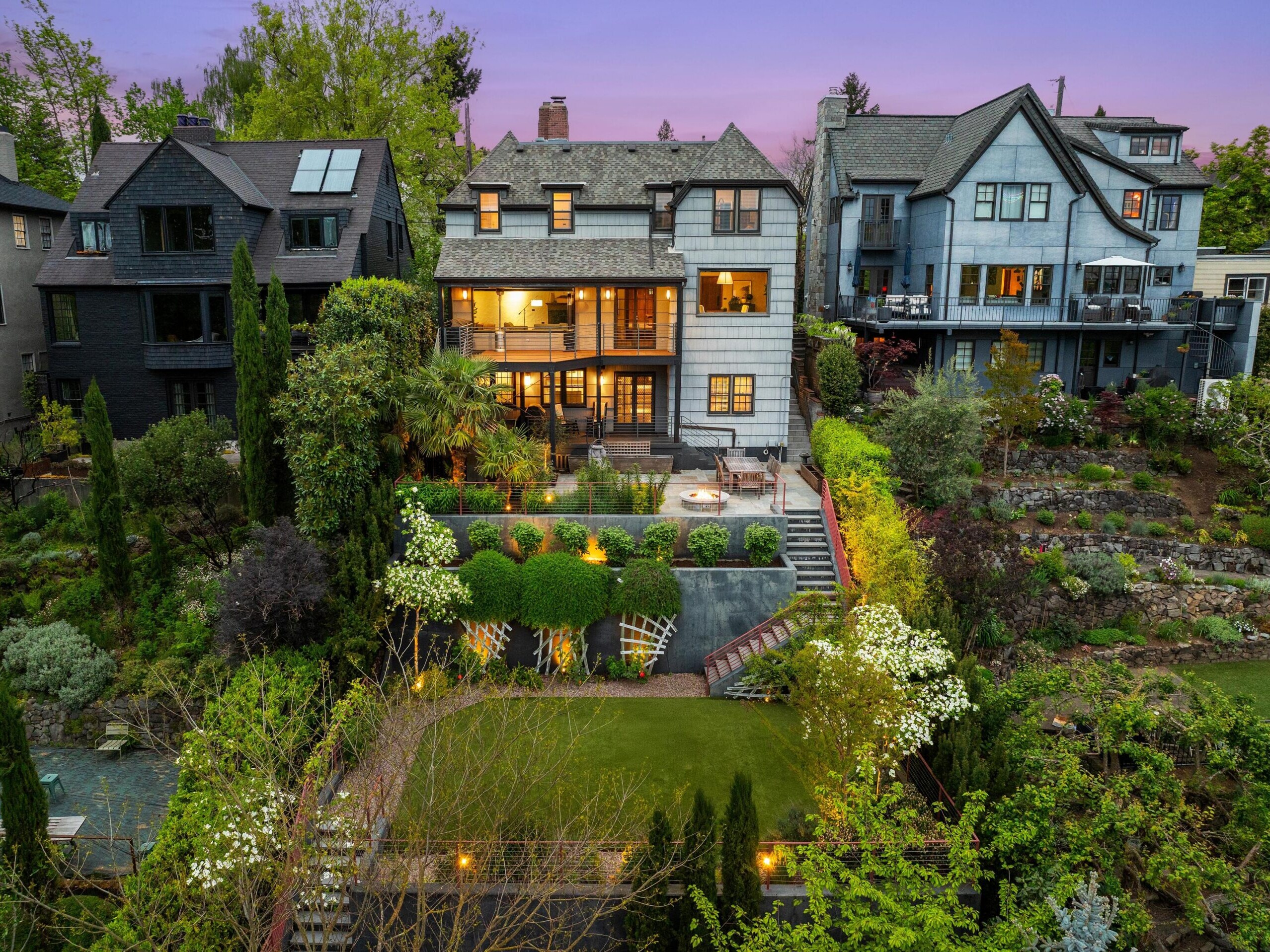 A large, elegant house with balconies and bright windows sits among lush gardens and trees at twilight, surrounded by other similar homes; a terraced yard and outdoor seating area are visible in the foreground.