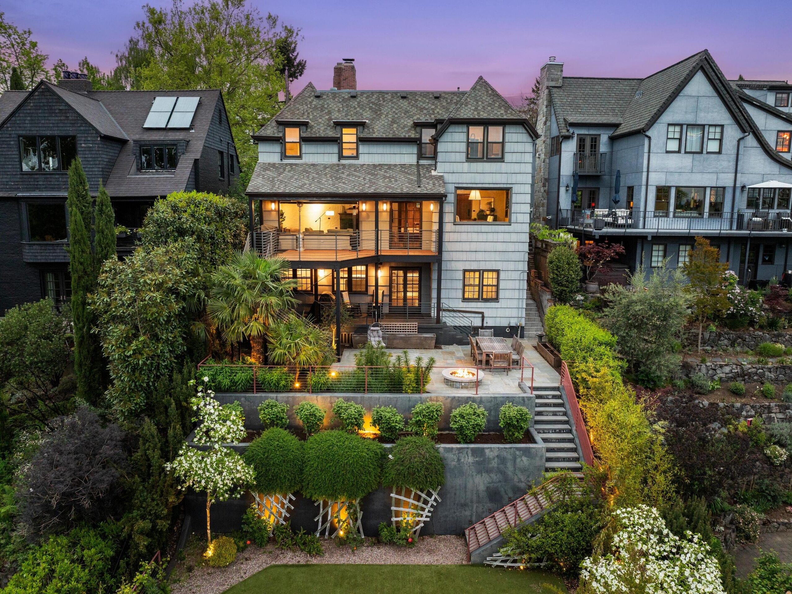 A large, three-story house with gray siding and lit windows sits on a landscaped slope, featuring multiple balconies, a patio with seating, lush greenery, and garden lights at dusk. Neighboring houses are visible on both sides.