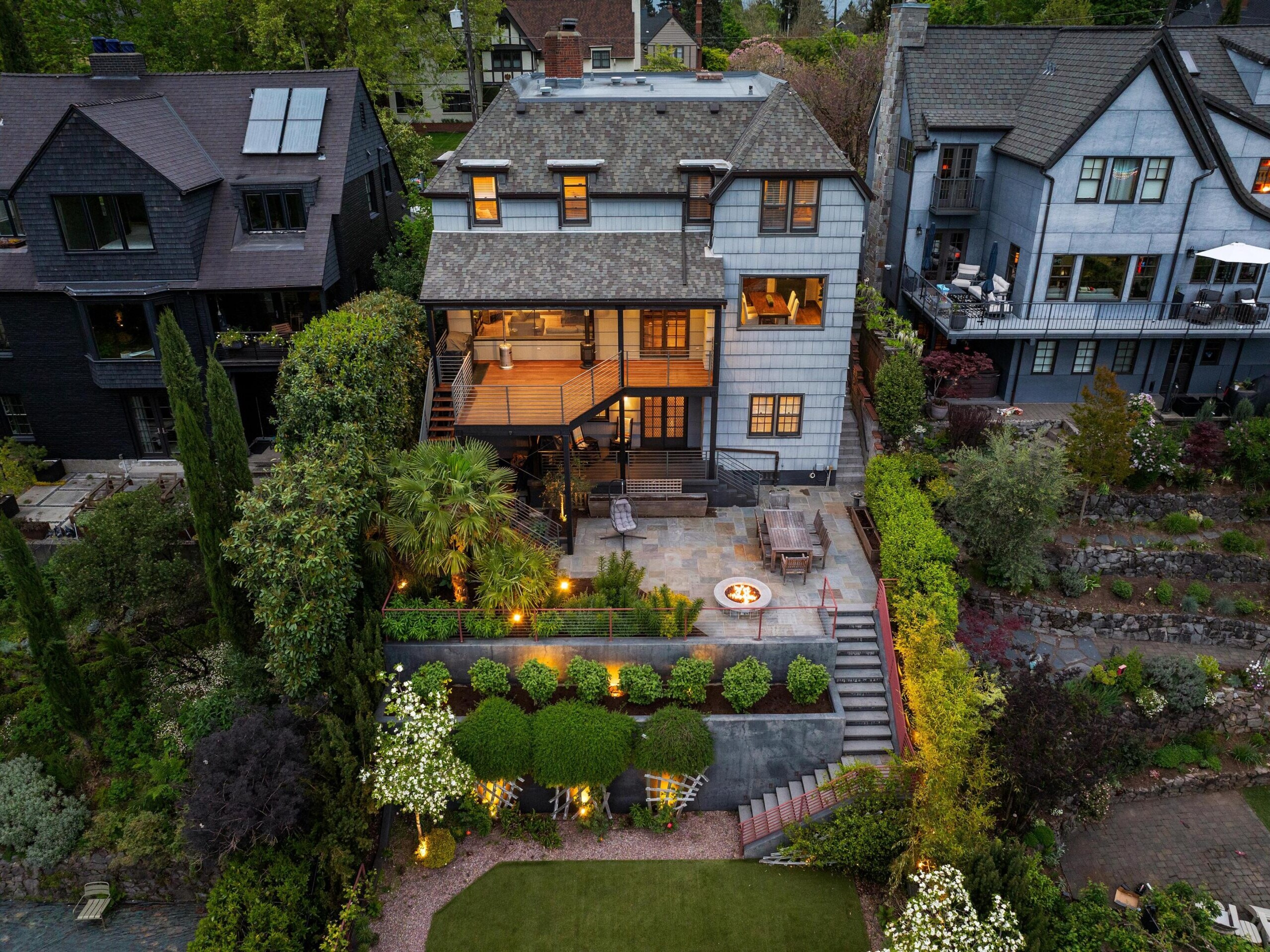 Aerial view of a multi-story house with large windows, a spacious balcony, and a landscaped backyard featuring a fire pit, outdoor seating, illuminated garden, stone paths, and lush greenery. Neighboring houses are visible on both sides.