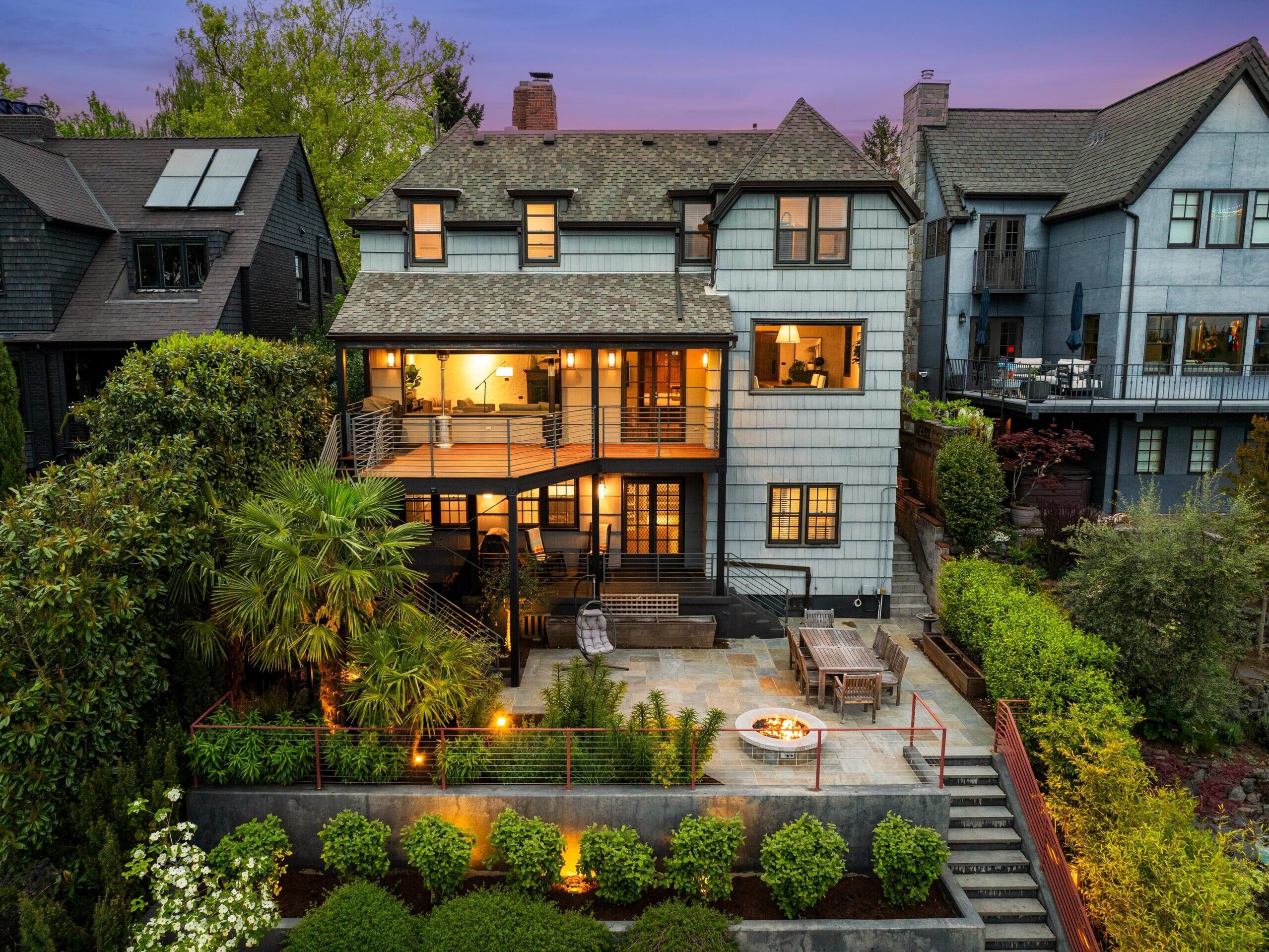 A large, modern house with multiple stories, warm lights glowing inside and on the patio, surrounded by lush greenery and outdoor seating areas, photographed at dusk.
