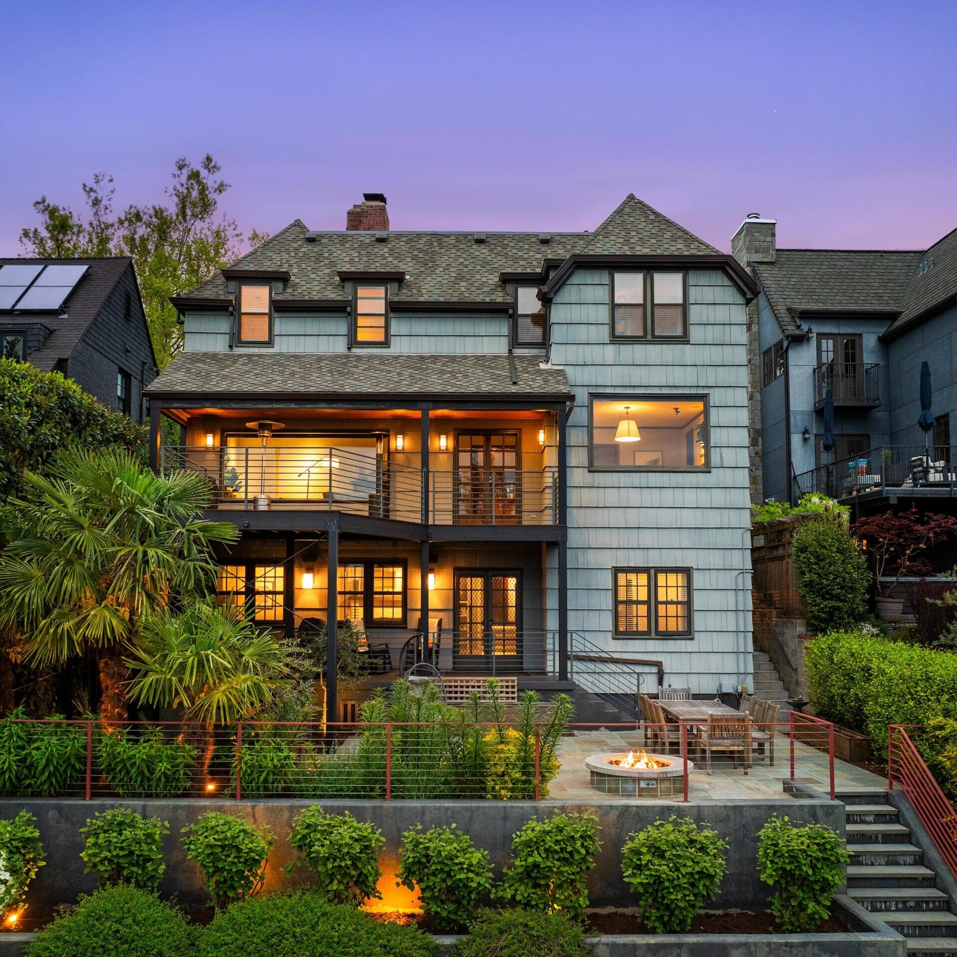 Three-story house with blue-gray siding, large windows, and balconies, warmly lit at dusk. Lush landscaping, palm trees, and a patio with a fire pit and seating area are in the foreground. Neighboring homes are visible.