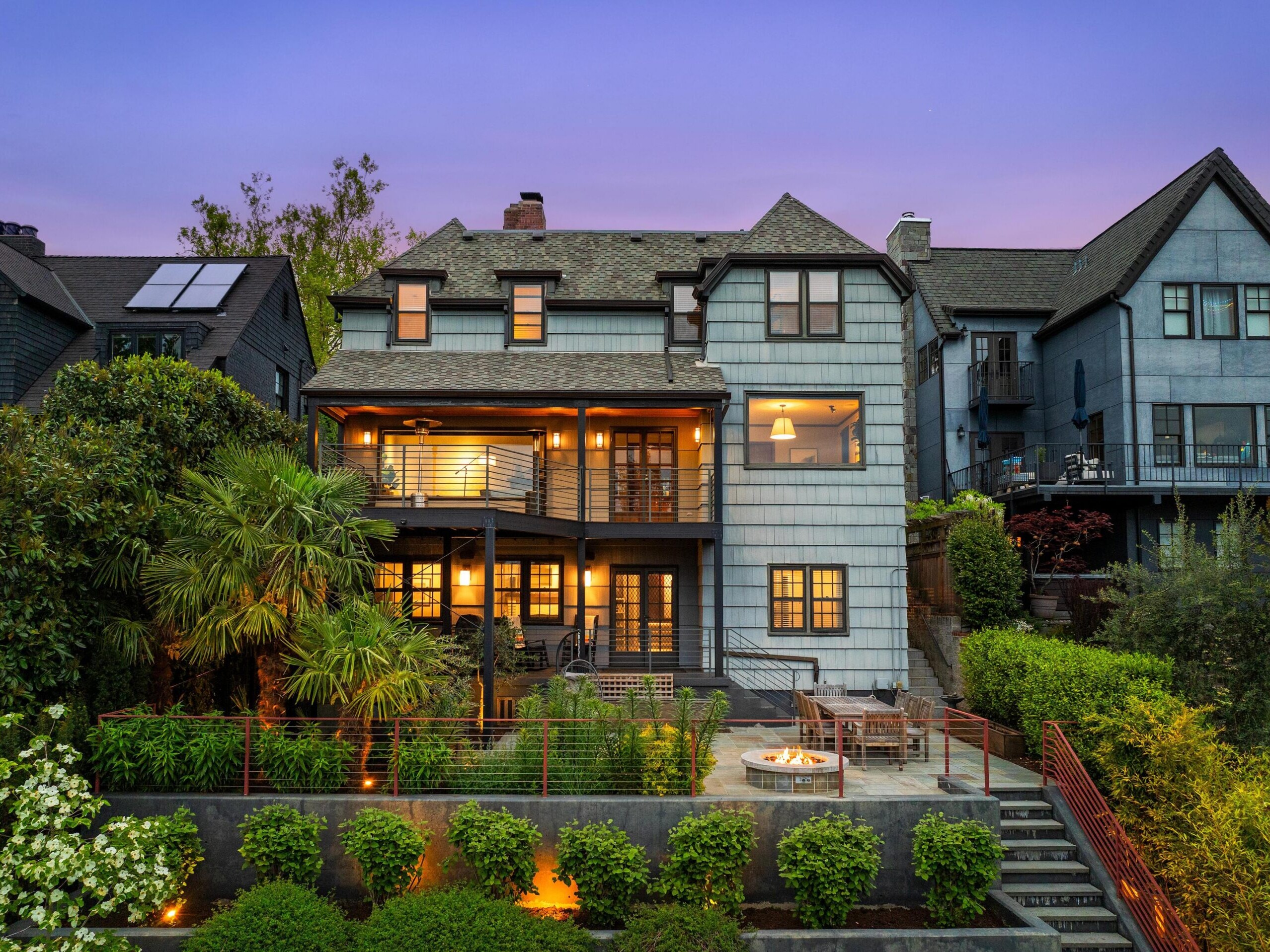 Three-story house with blue-gray siding, large windows, and balconies, warmly lit at dusk. Lush landscaping, palm trees, and a patio with a fire pit and seating area are in the foreground. Neighboring homes are visible.
