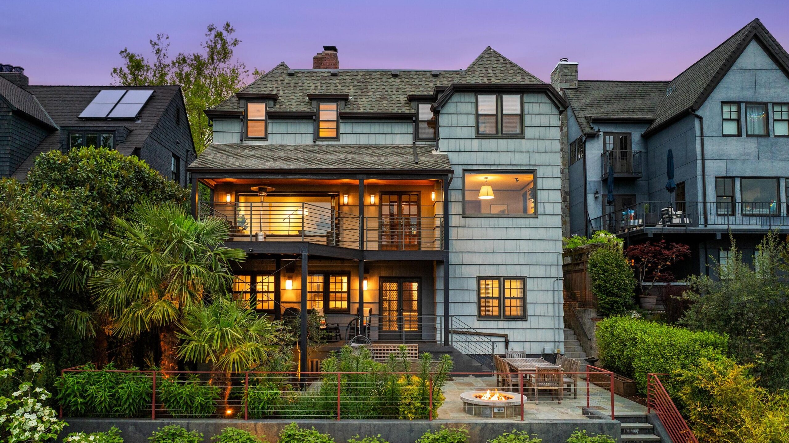 Three-story house with blue-gray siding, large windows, and balconies, warmly lit at dusk. Lush landscaping, palm trees, and a patio with a fire pit and seating area are in the foreground. Neighboring homes are visible.