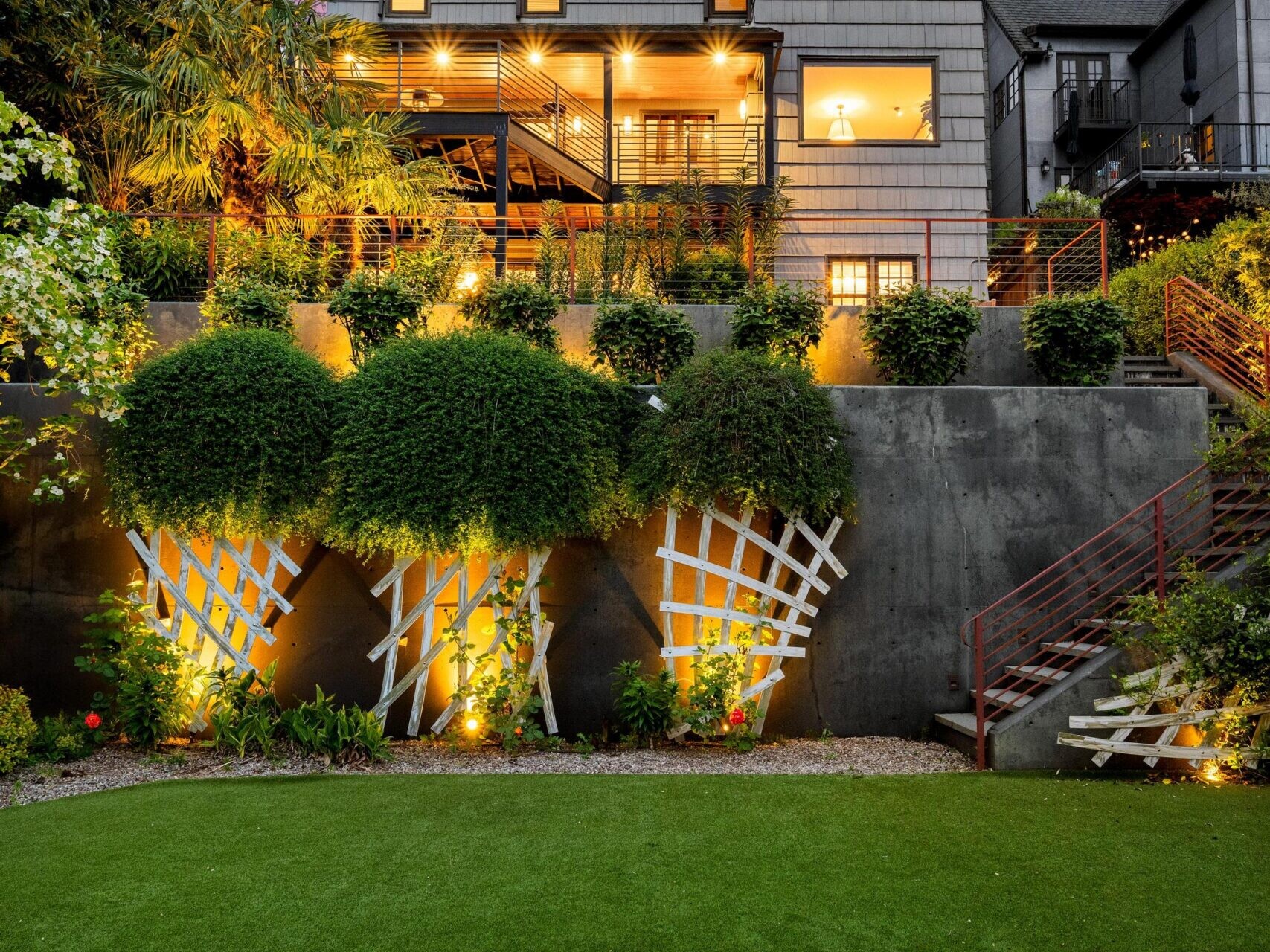A modern house with large windows and warm exterior lights is set above a lush, green lawn. Decorative trellises with climbing plants and outdoor lighting accent the retaining wall in the landscaped backyard at dusk.