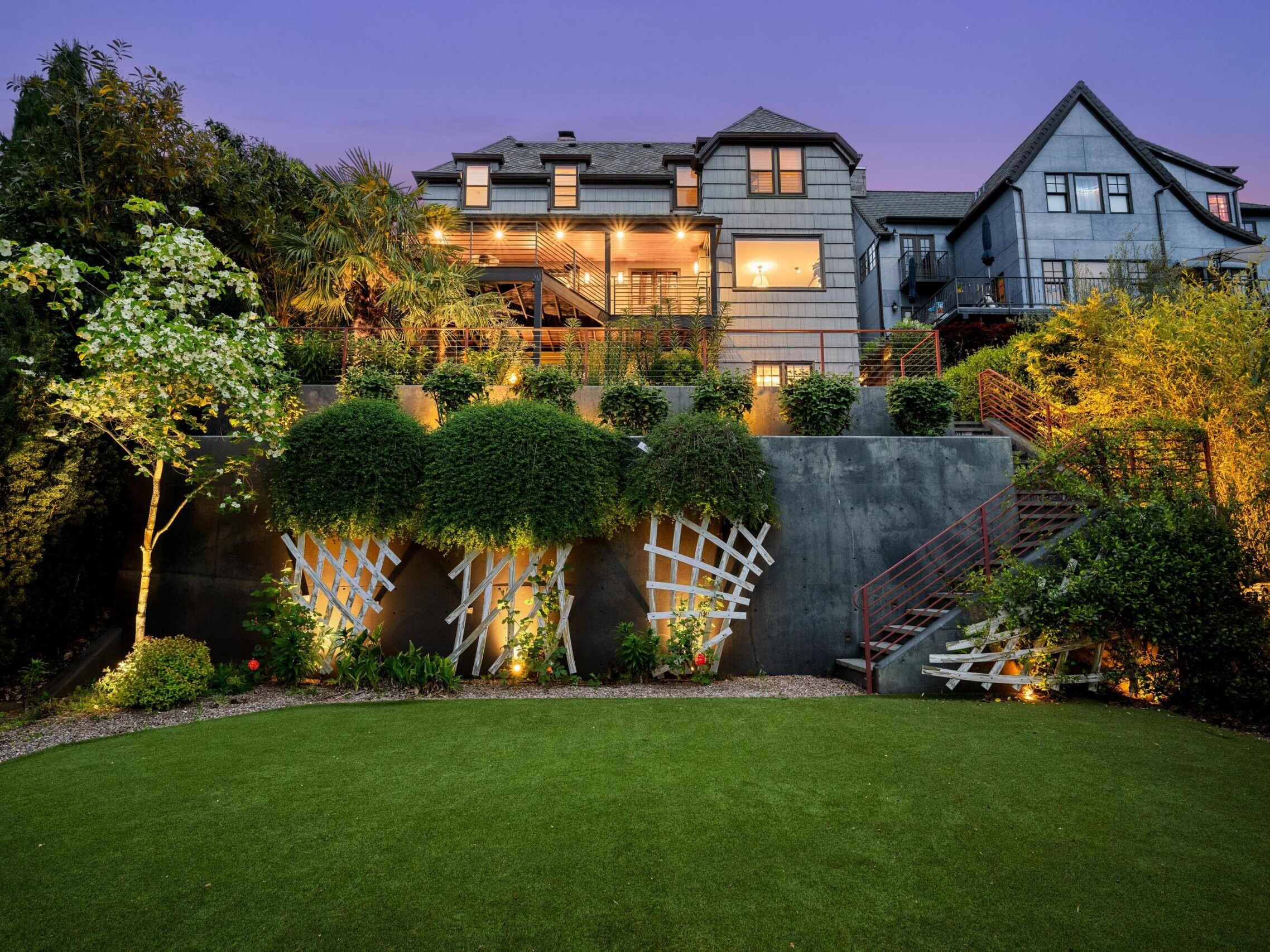 A large, modern house with multiple stories and balconies is illuminated at dusk. The backyard features a manicured lawn, terraced gardens, decorative trellises, and accent lighting among lush greenery.