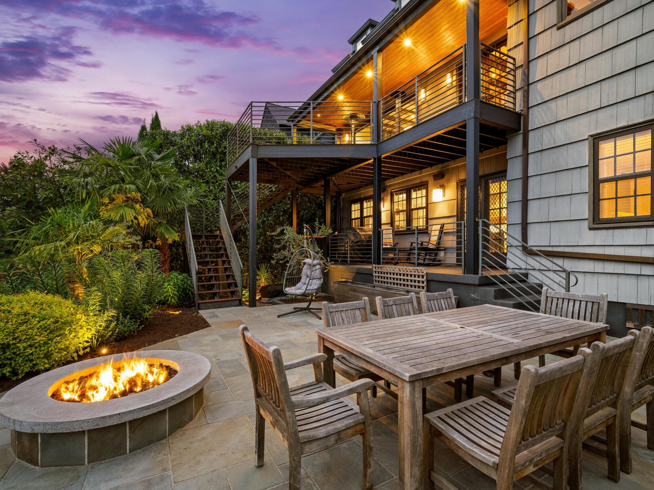 Two-story house with a covered balcony and patio, wooden outdoor dining set, circular fire pit, lush landscaping, and a sunset sky in the background.