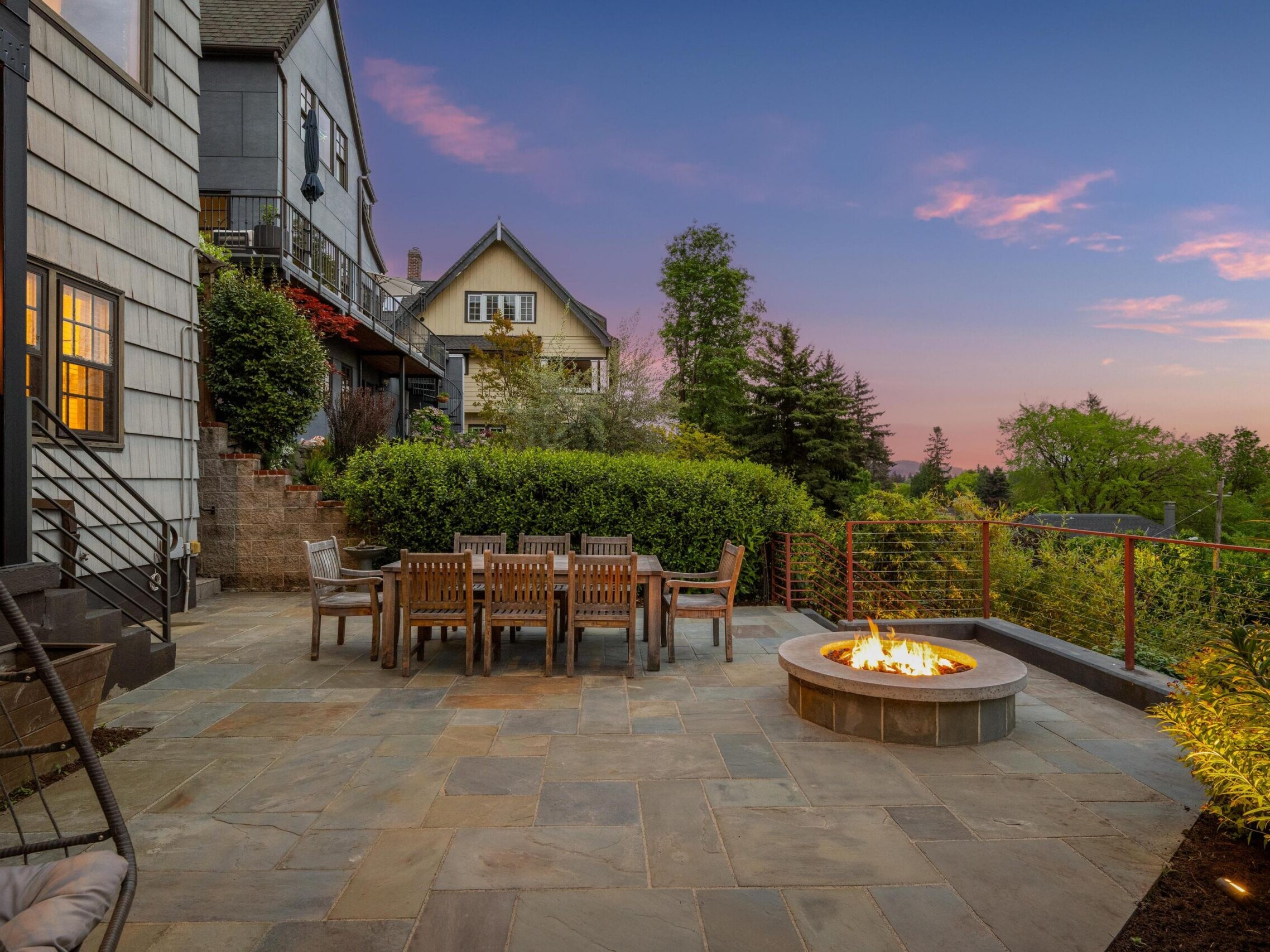 A spacious stone patio with a dining table and chairs sits beside a round fire pit. Lush greenery and houses surround the area under a colorful sunset sky.