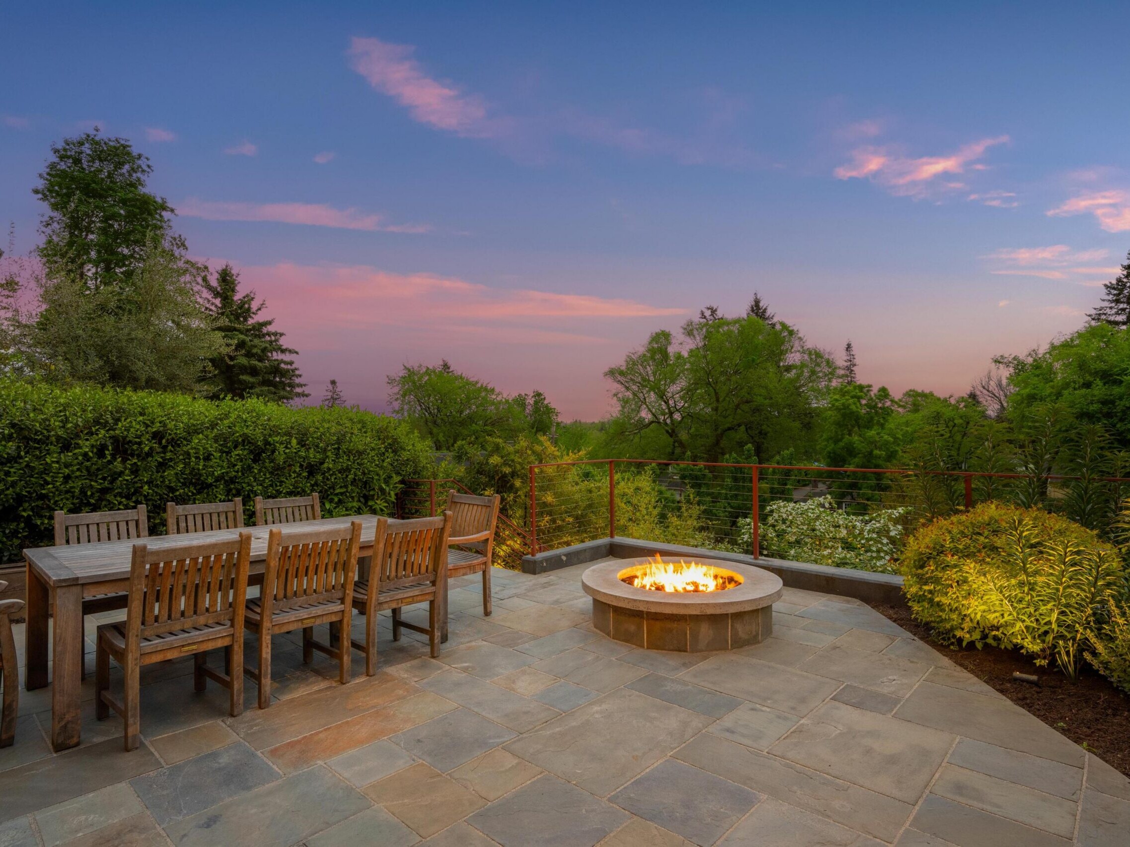 A spacious patio with a long wooden dining table and chairs sits beside a lit fire pit, overlooking lush greenery and trees under a colorful sunset sky.