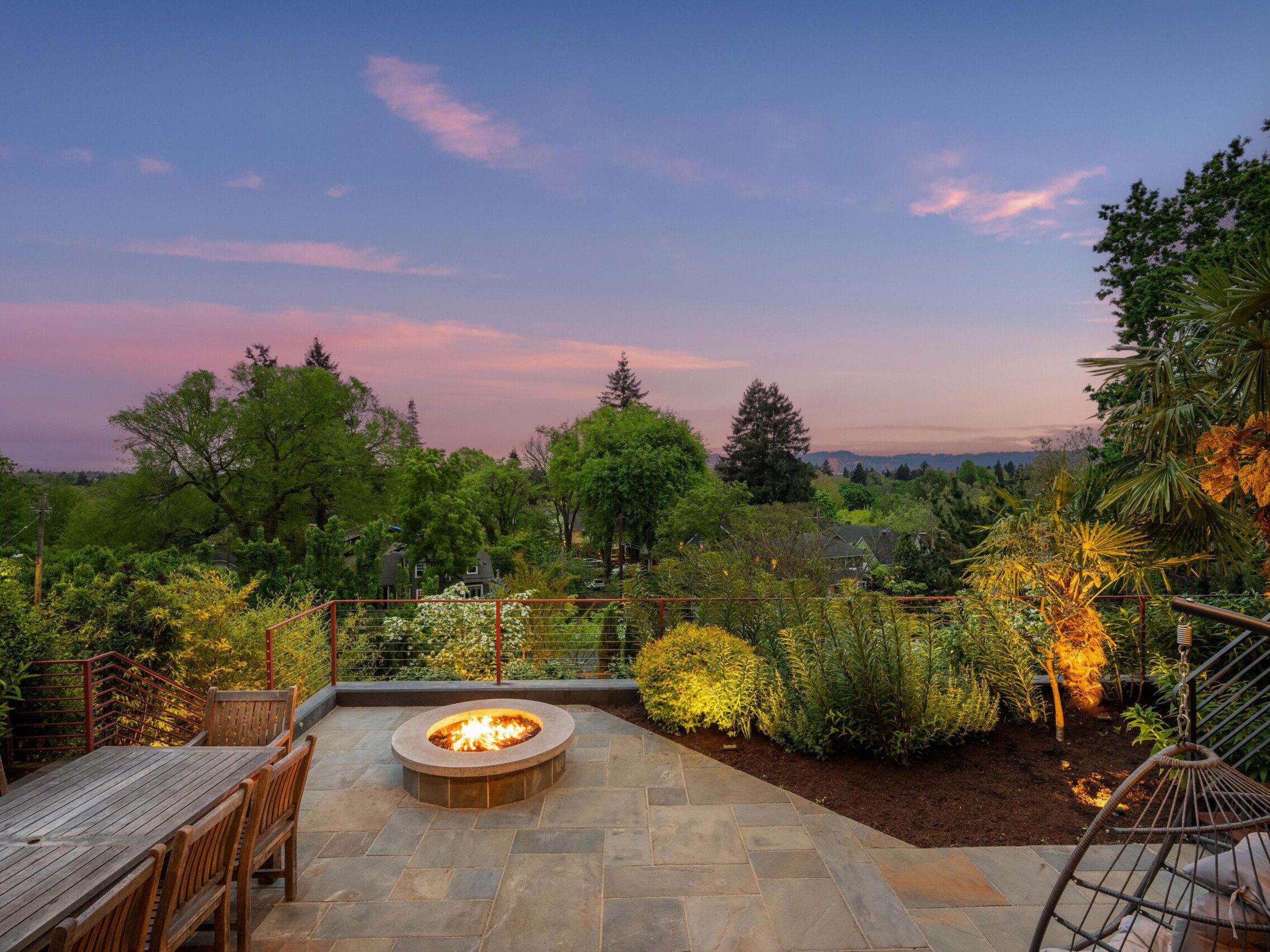A stone patio with a firepit, wooden outdoor dining table, and lush greenery, overlooking a scenic landscape at sunset with a pink and purple sky.