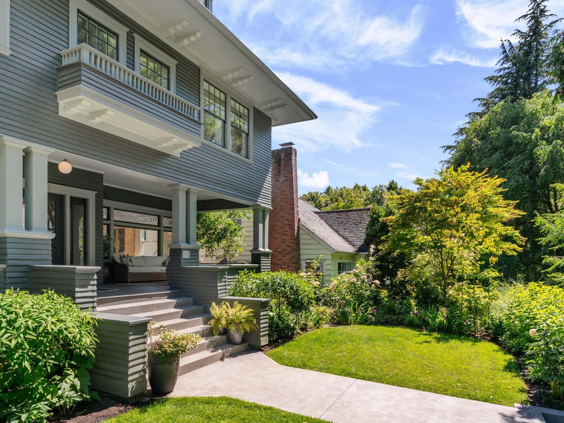 A beautiful gray two-story house with a covered front porch, white trim, and lush green landscaping on a sunny day with a blue sky and scattered clouds.