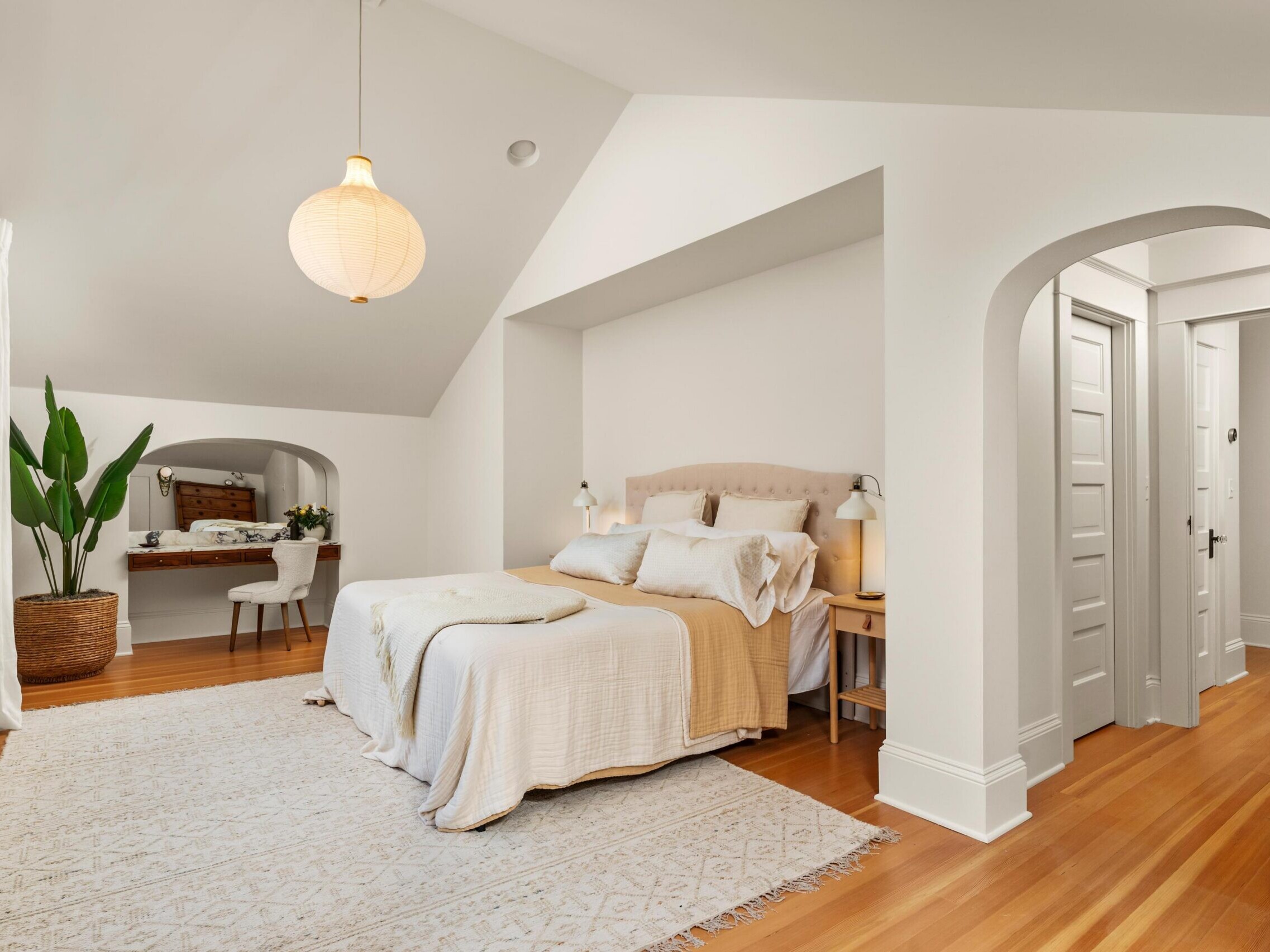 Bright bedroom with a large bed, neutral bedding, bedside lamps, a potted plant, wooden floors, a white rug, a pendant light, and a cozy nook with a desk and chair by the window.