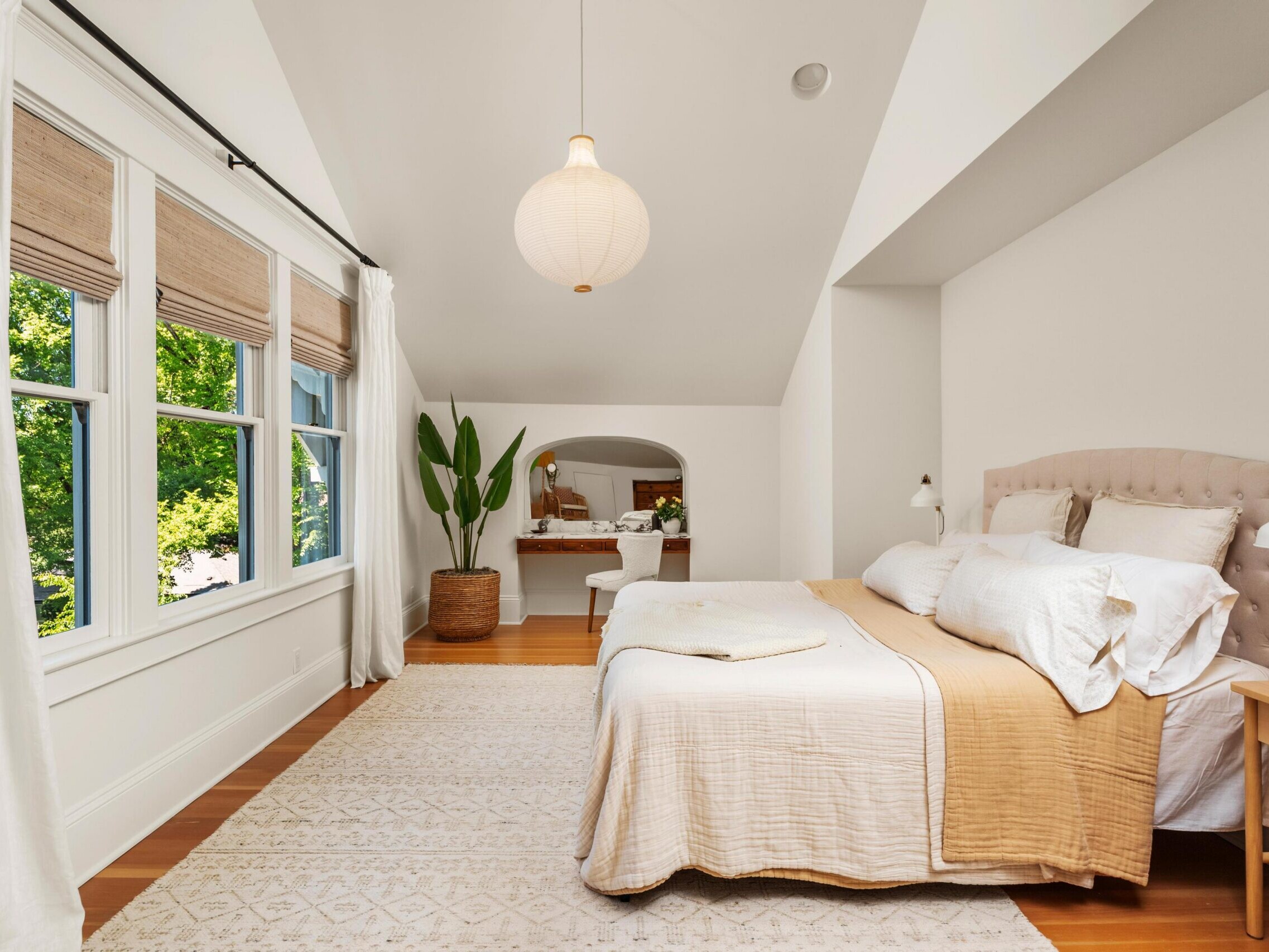 Bright bedroom with large windows, light-colored walls, a beige bed with white pillows, a light rug, wooden floors, a potted plant, and a small work desk in the background under an archway.