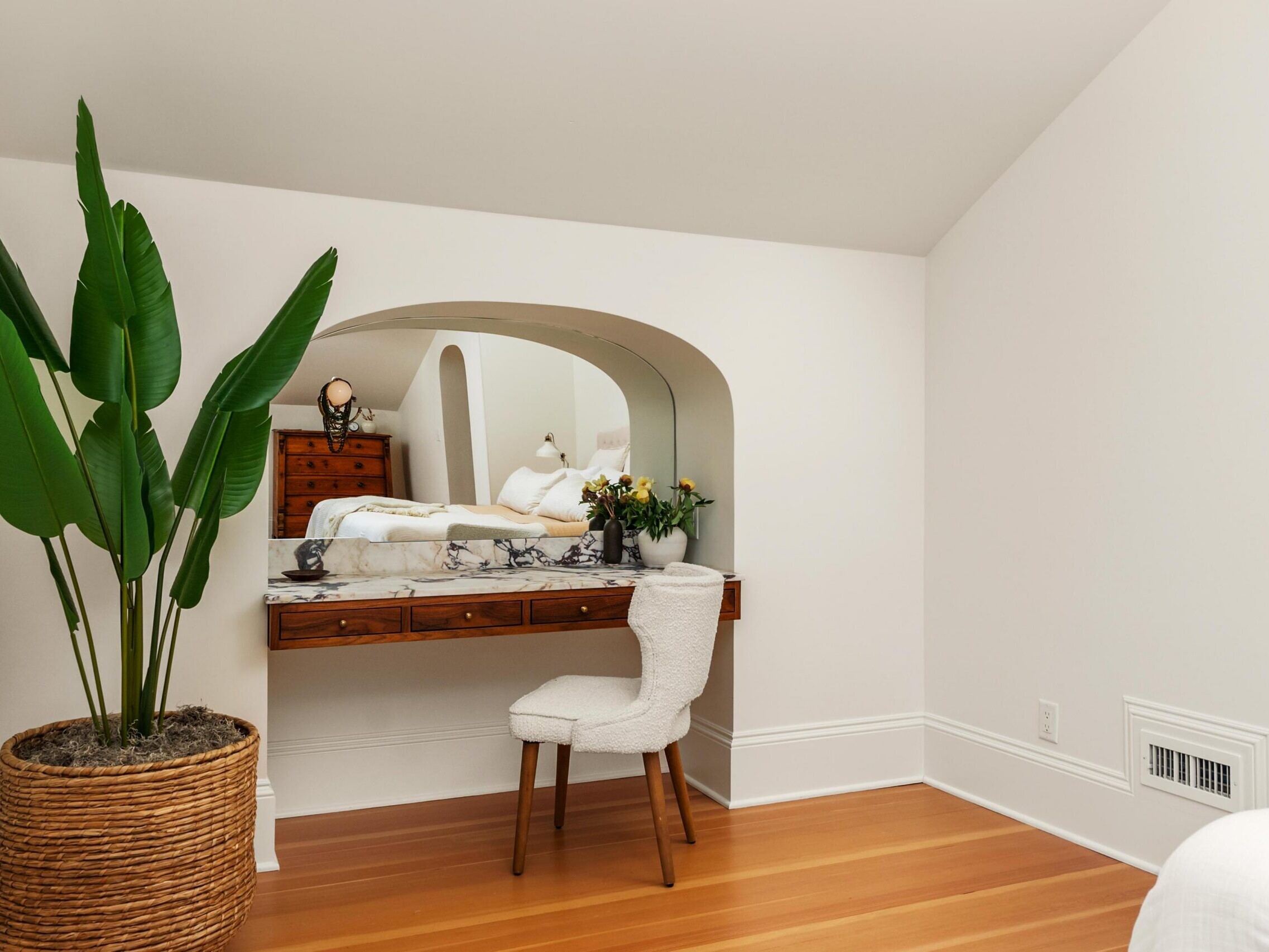 A cozy bedroom corner with a wooden vanity table topped with flowers and decor, a cushioned chair, a large potted plant, and a mirror reflecting a neatly made bed in a softly lit, white-walled room with wood flooring.