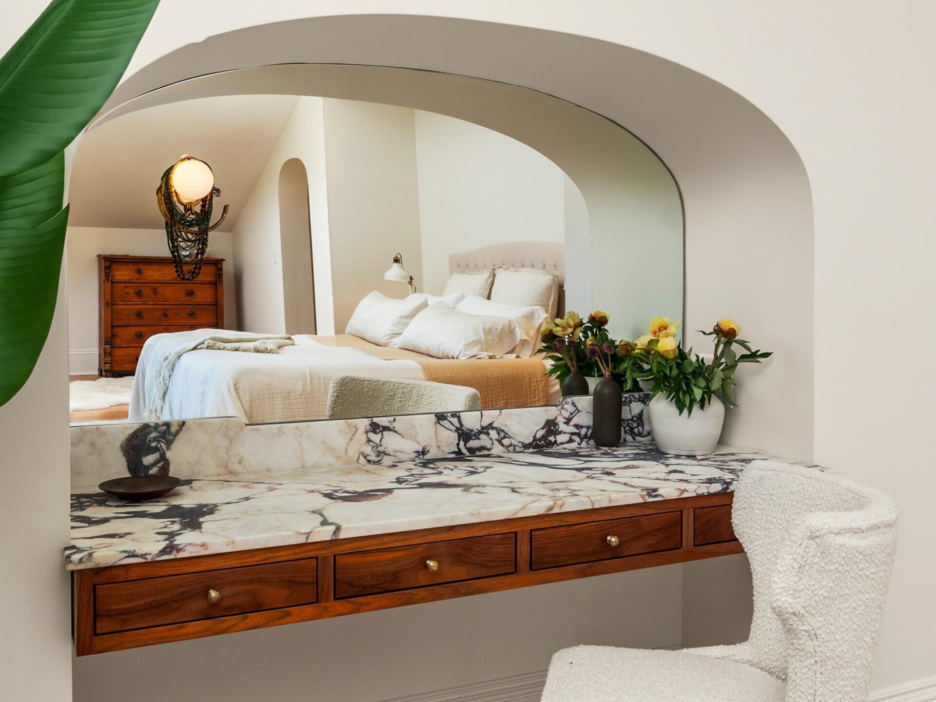 A cozy vanity area with a marble countertop, wooden drawers, and a plush white chair sits beneath an arched opening, reflecting a neatly made bed and dresser in the adjoining bedroom. Fresh flowers decorate the vanity.
